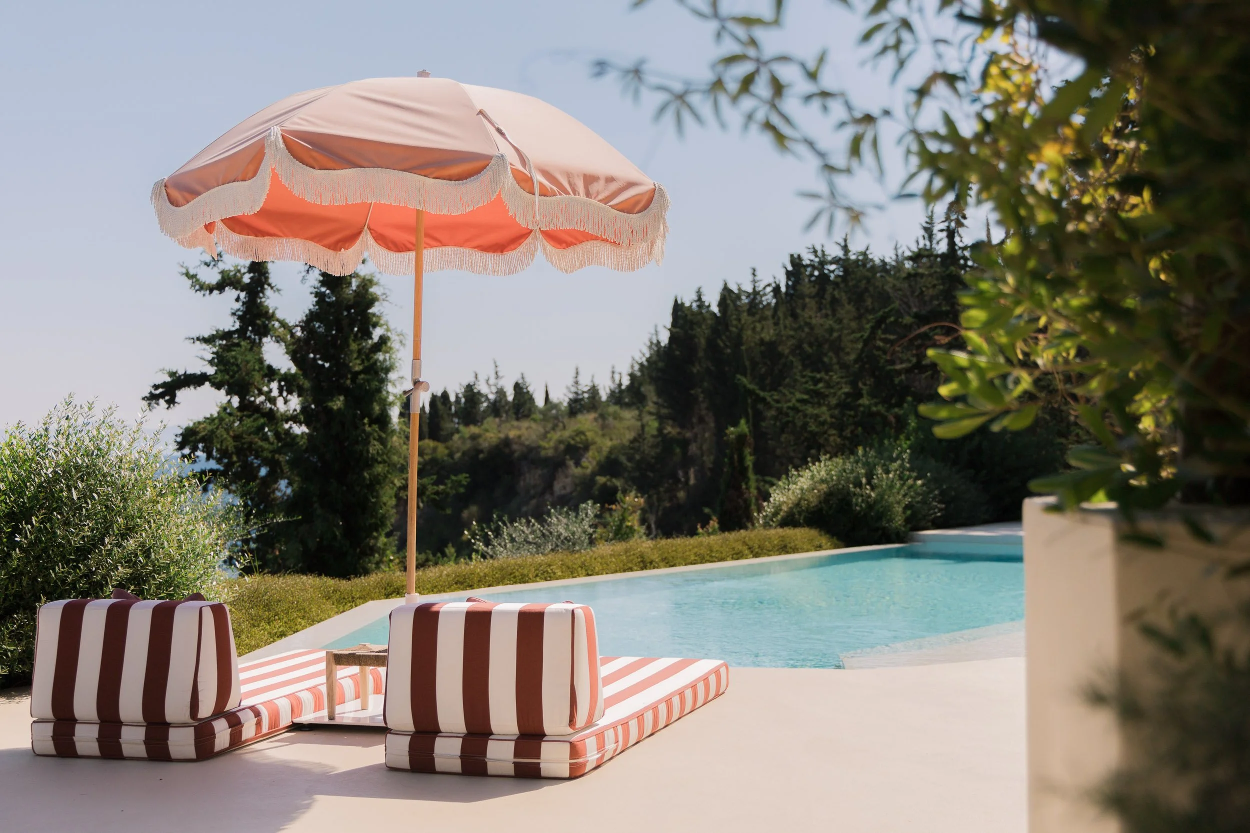 A swimming pool with striped lounge chairs and a pink patio umbrella with fringe, surrounded by greenery and trees in the background.