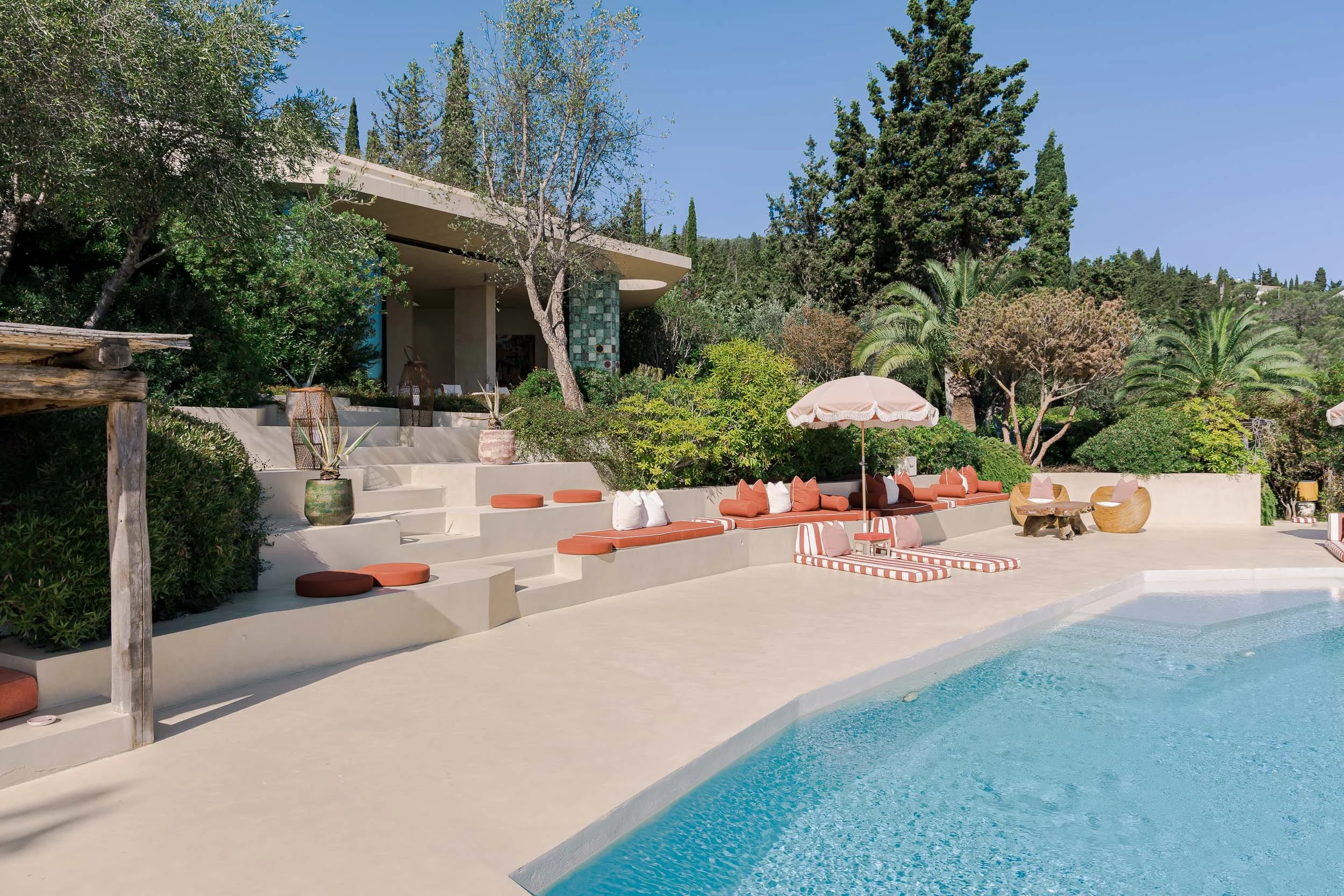 Poolside seating area with orange cushions, striped umbrellas, and lush green trees in the background.
