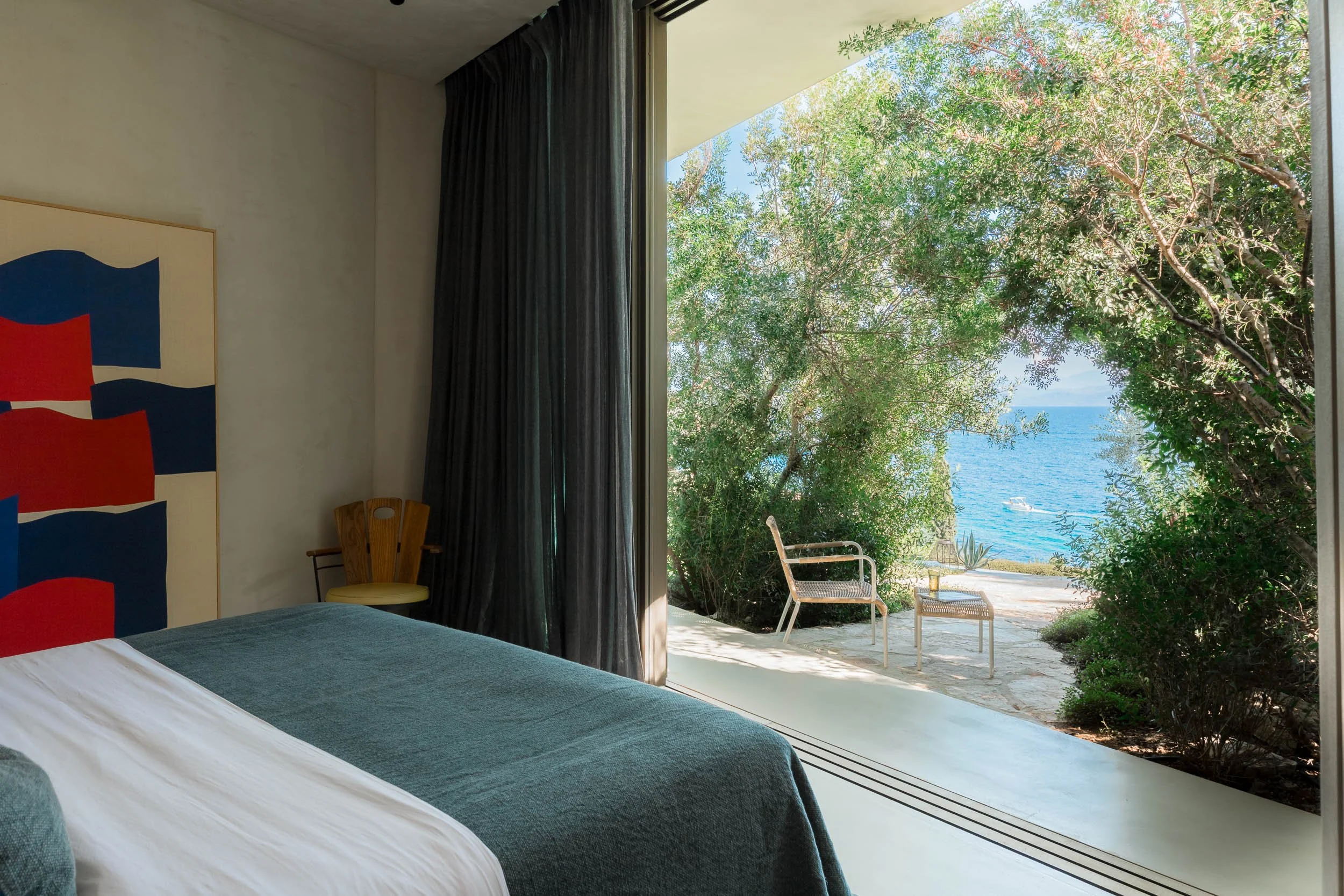 A bedroom with a large open window showing a view of trees, the ocean, and boats. Inside, there is a bed with white sheets and a teal blanket, a wooden chair, and dark curtains.