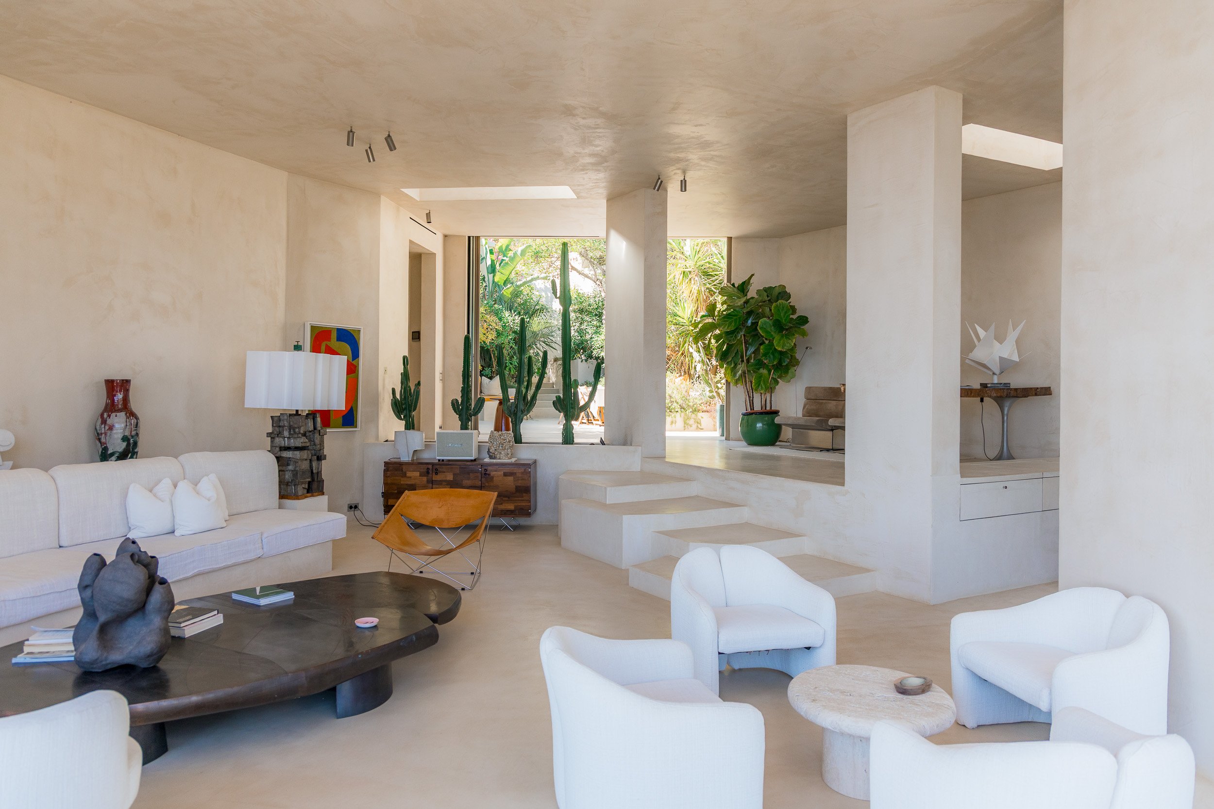 Modern living room with white furniture, green plants, and large windows.
