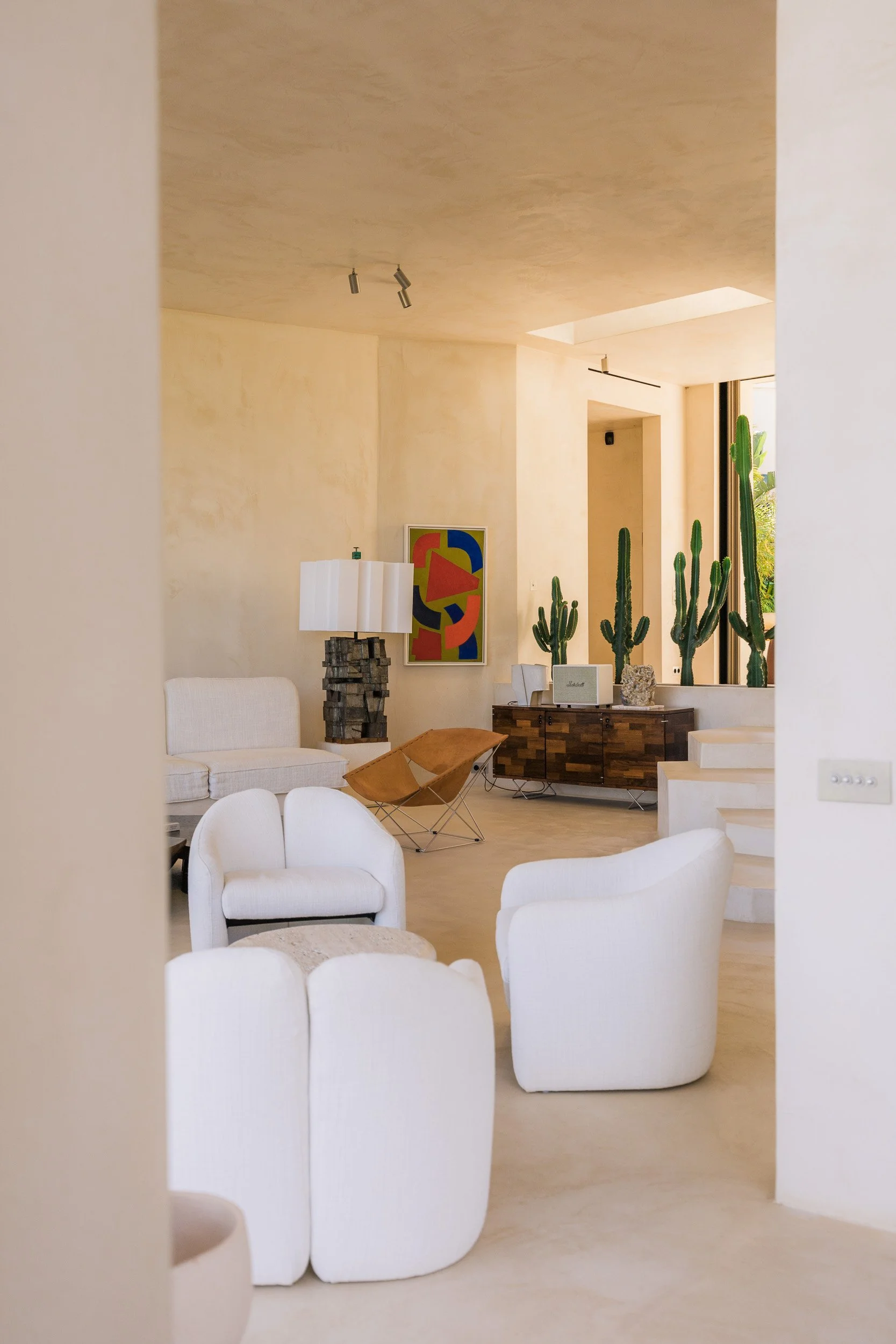 Living room with white chairs, beige sofa, wooden cabinet, cactus plants in the background, abstract colorful artwork on the wall, and a sunlit window.