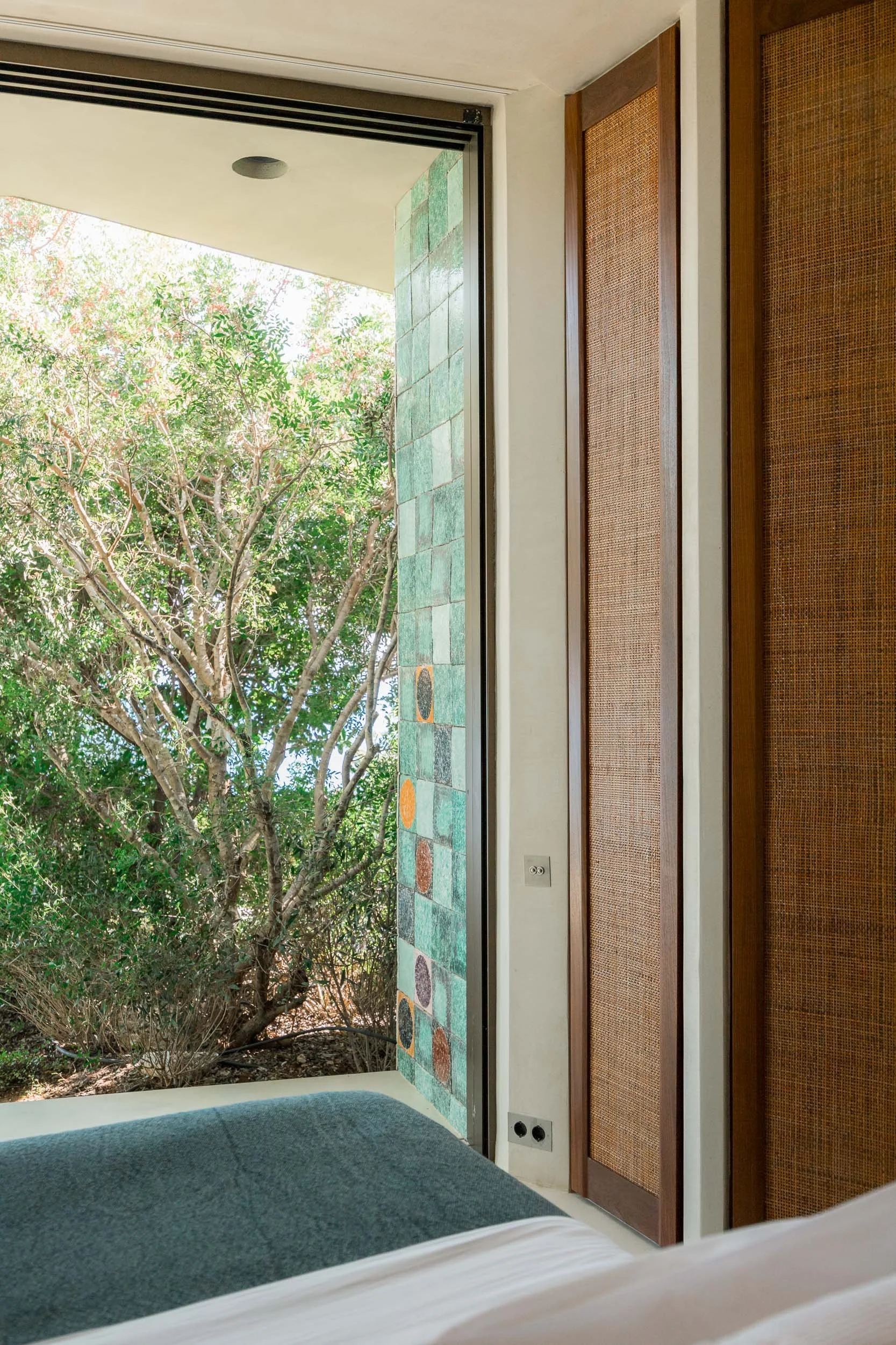 Photo of a bedroom with a sliding glass door opening to a view of trees outside. The room has a bed with white sheets and a dark gray blanket, and a colorful tiled wall portion adjacent to the door. There are wooden cabinets with woven rattan fronts 