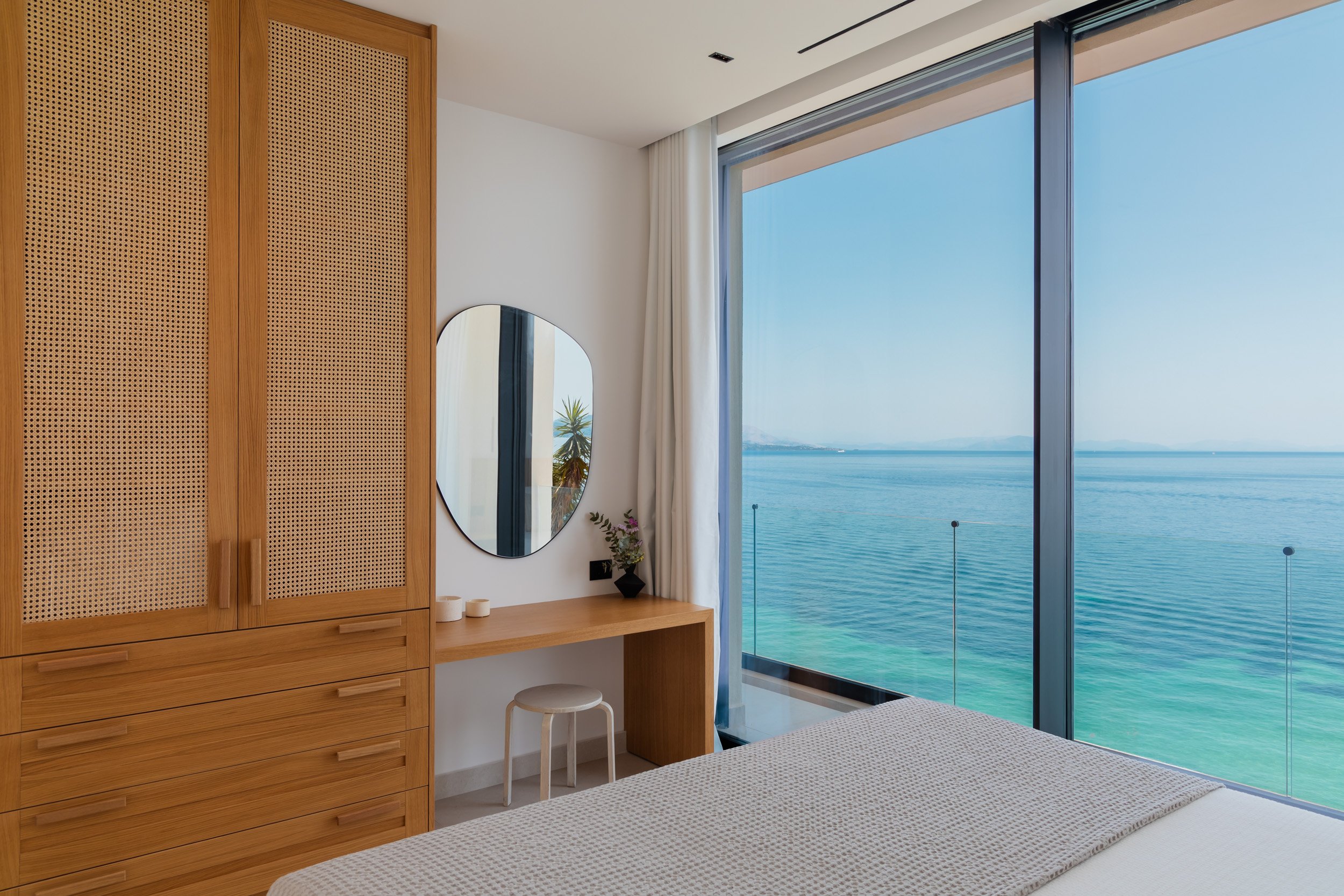 A bedroom with a large glass window overlooking a body of water, with a wooden wardrobe, a desk with a round mirror, and a stool. The bed is partially visible in the foreground.