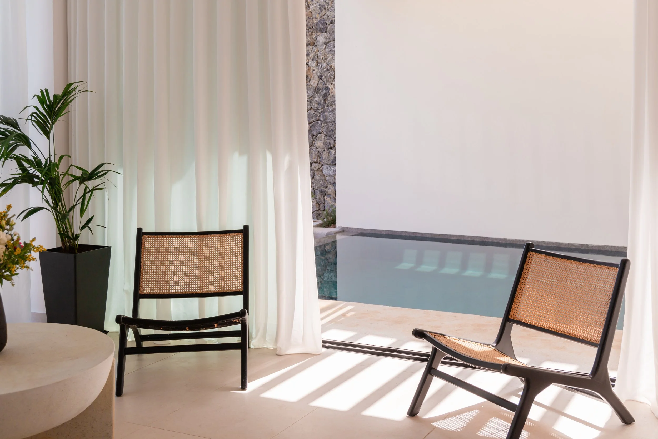Indoor area with two black chairs with woven seats, a large potted plant, and floor-length white curtains. Outside, a pool is visible with a white wall and stone wall in the background.