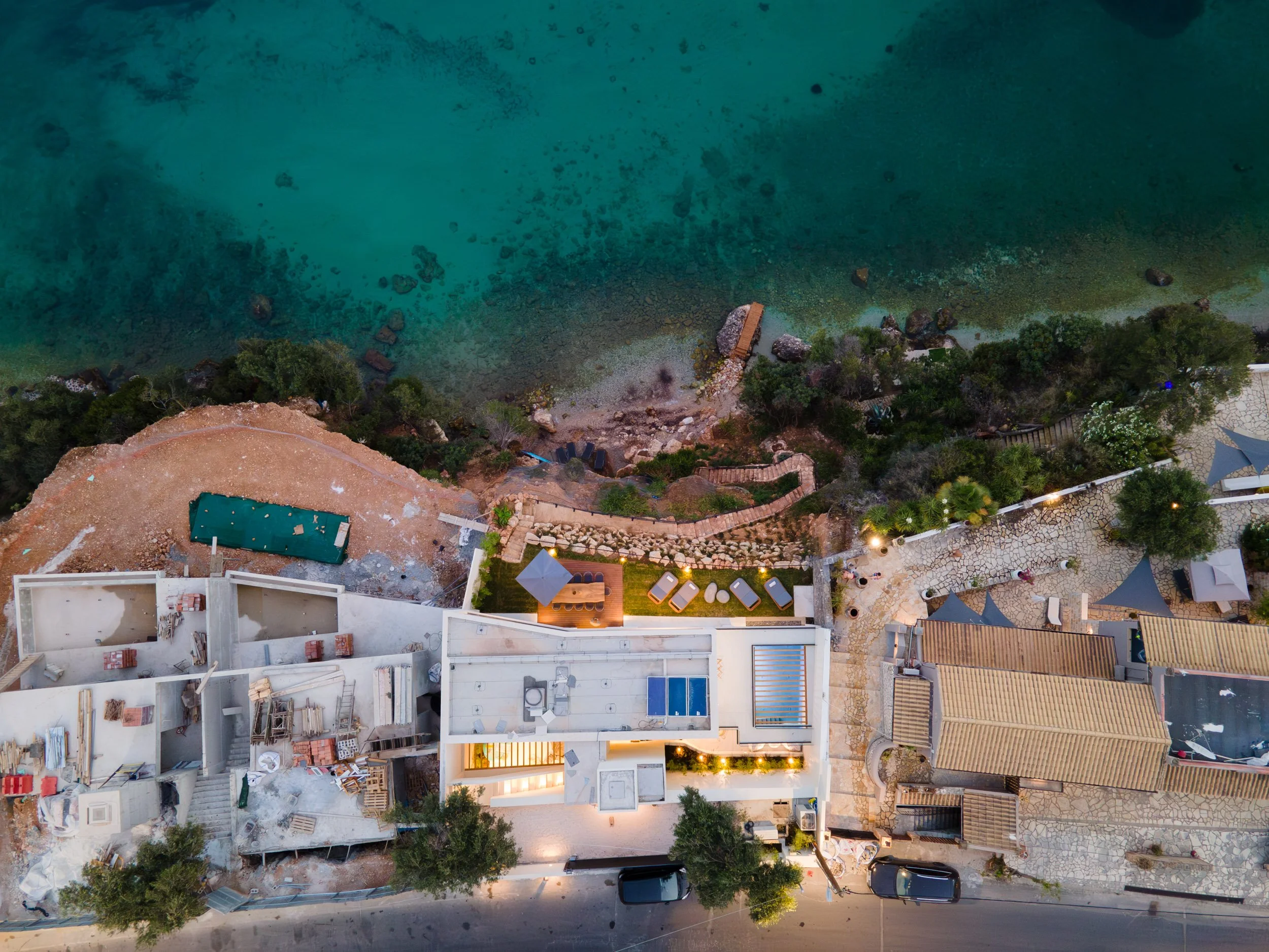 Aerial view of a coastal building under construction with nearby outdoor seating area and pathway, adjacent to a turquoise ocean with rocks and greenery.