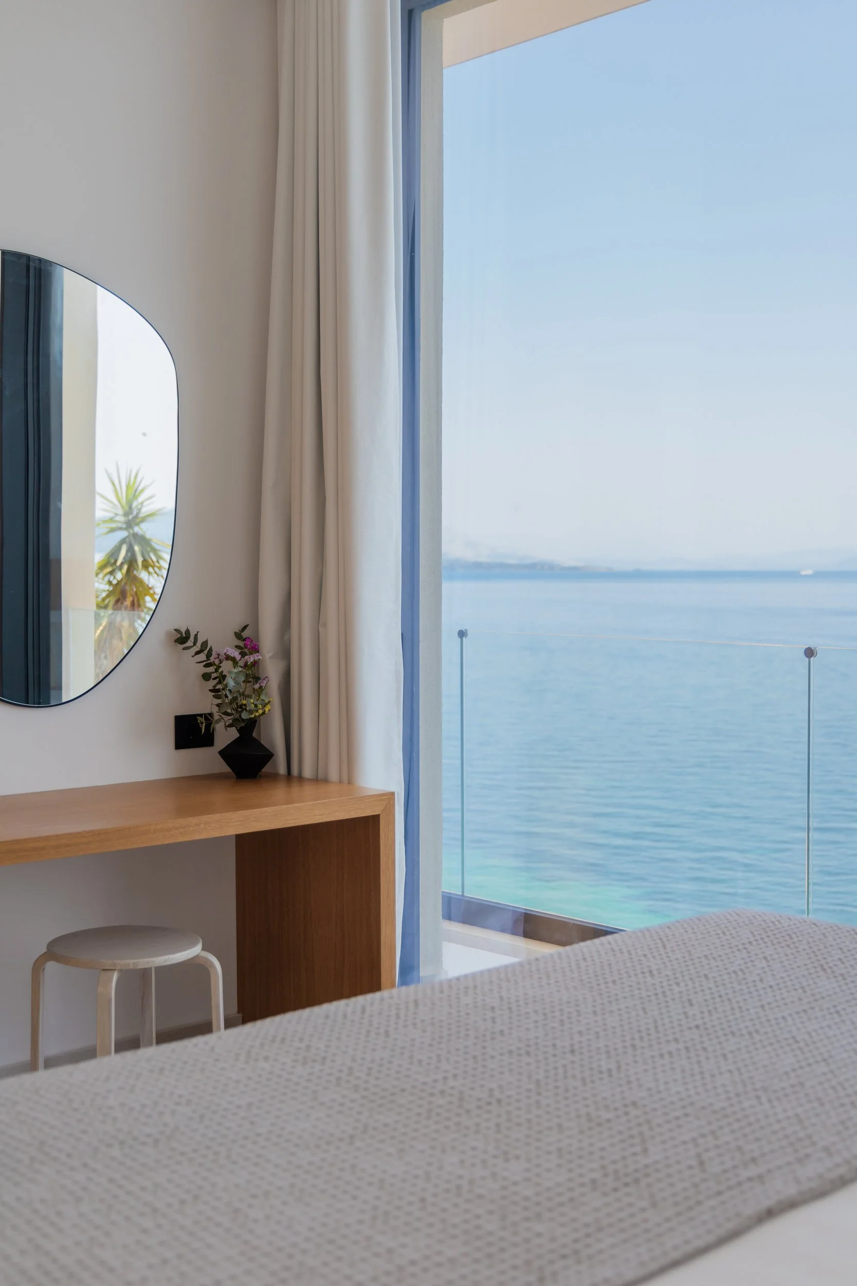A bedroom with a large window showing a body of water outside. The room has a wooden desk with a vase of flowers, a round mirror on the wall, a small white stool, and a bed with a beige blanket.