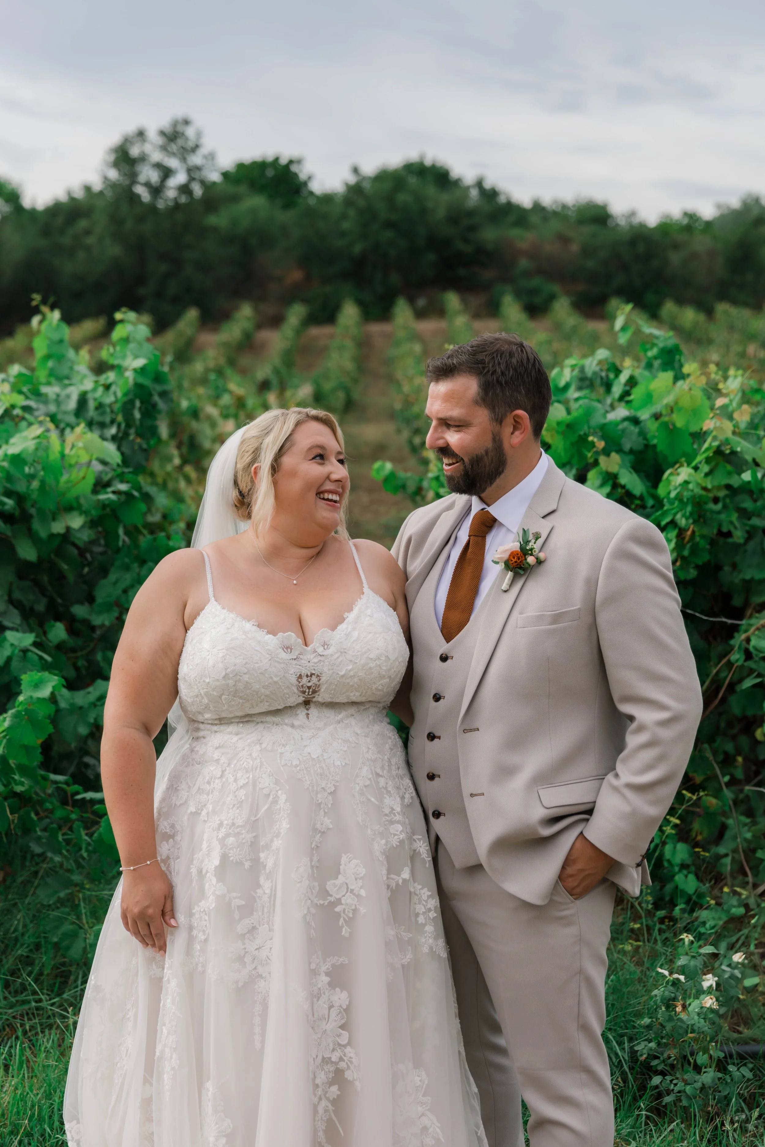 A bride and groom smiling at each other outdoors, surrounded by vineyard greenery and rows of grapevines.