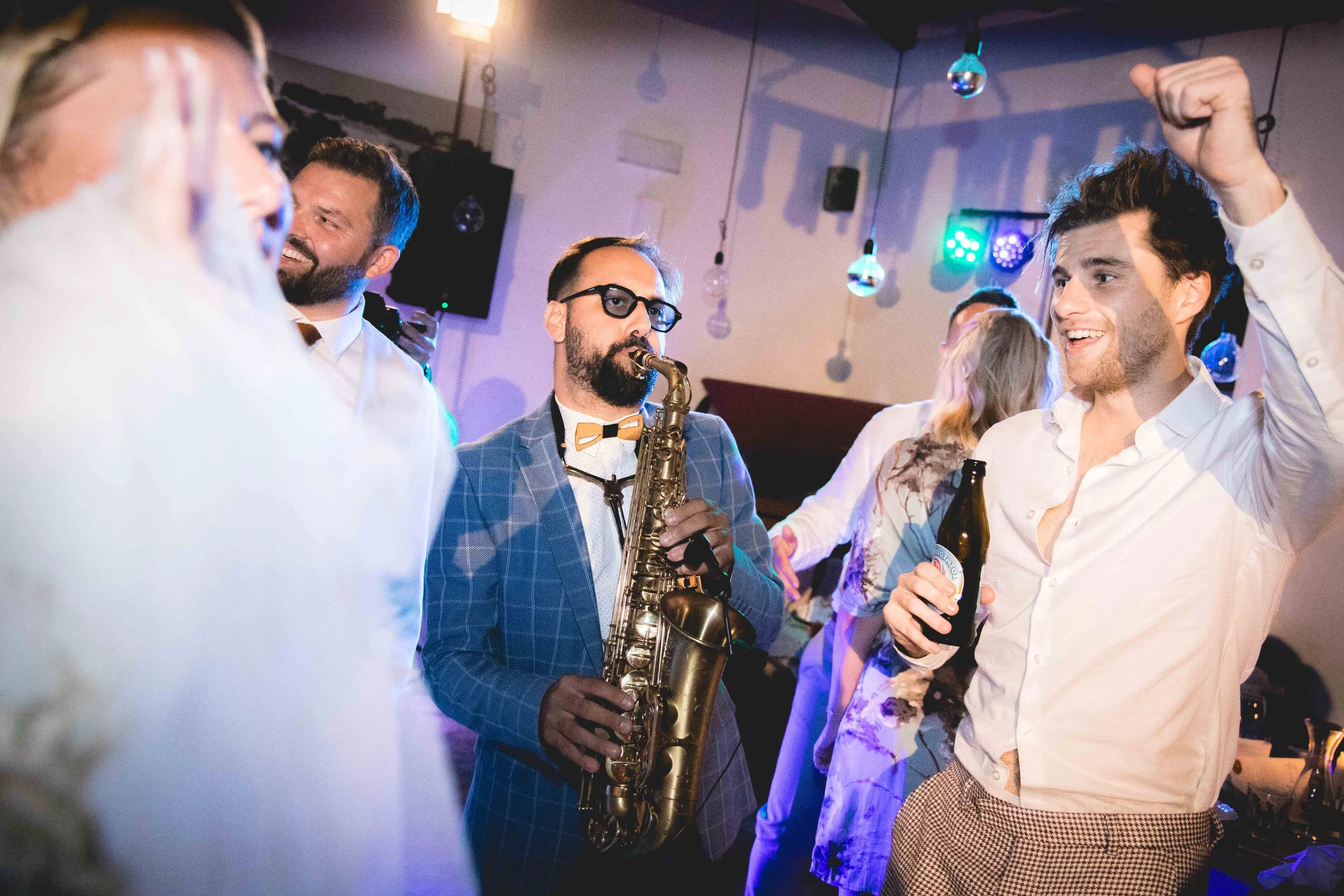 People dancing and enjoying music at a party, with a saxophonist playing in the center and colorful lights overhead.