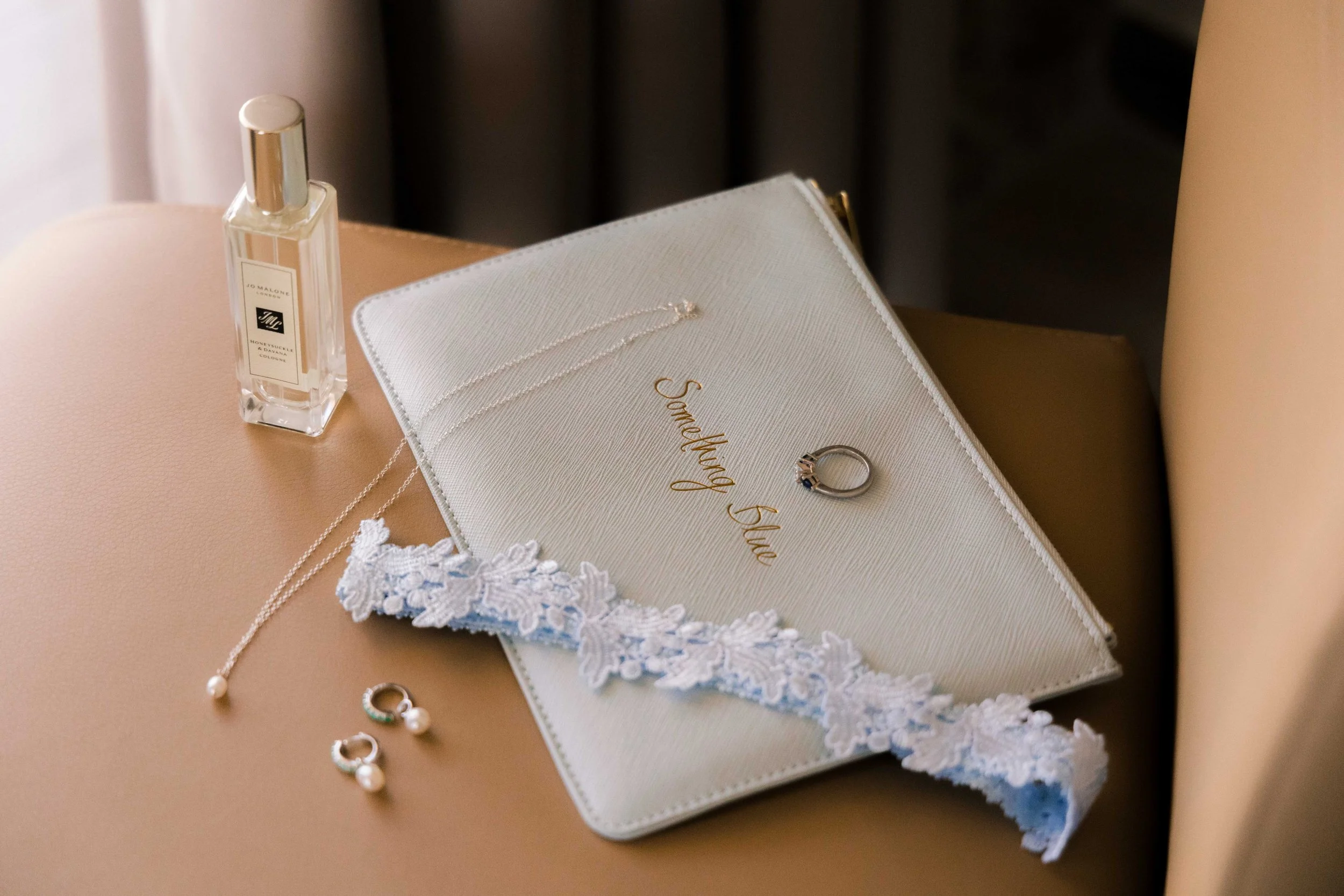 Wedding ring, lace garter, pearl necklace, pearl earrings, perfume bottle on a peach-colored surface with a beige chair back.