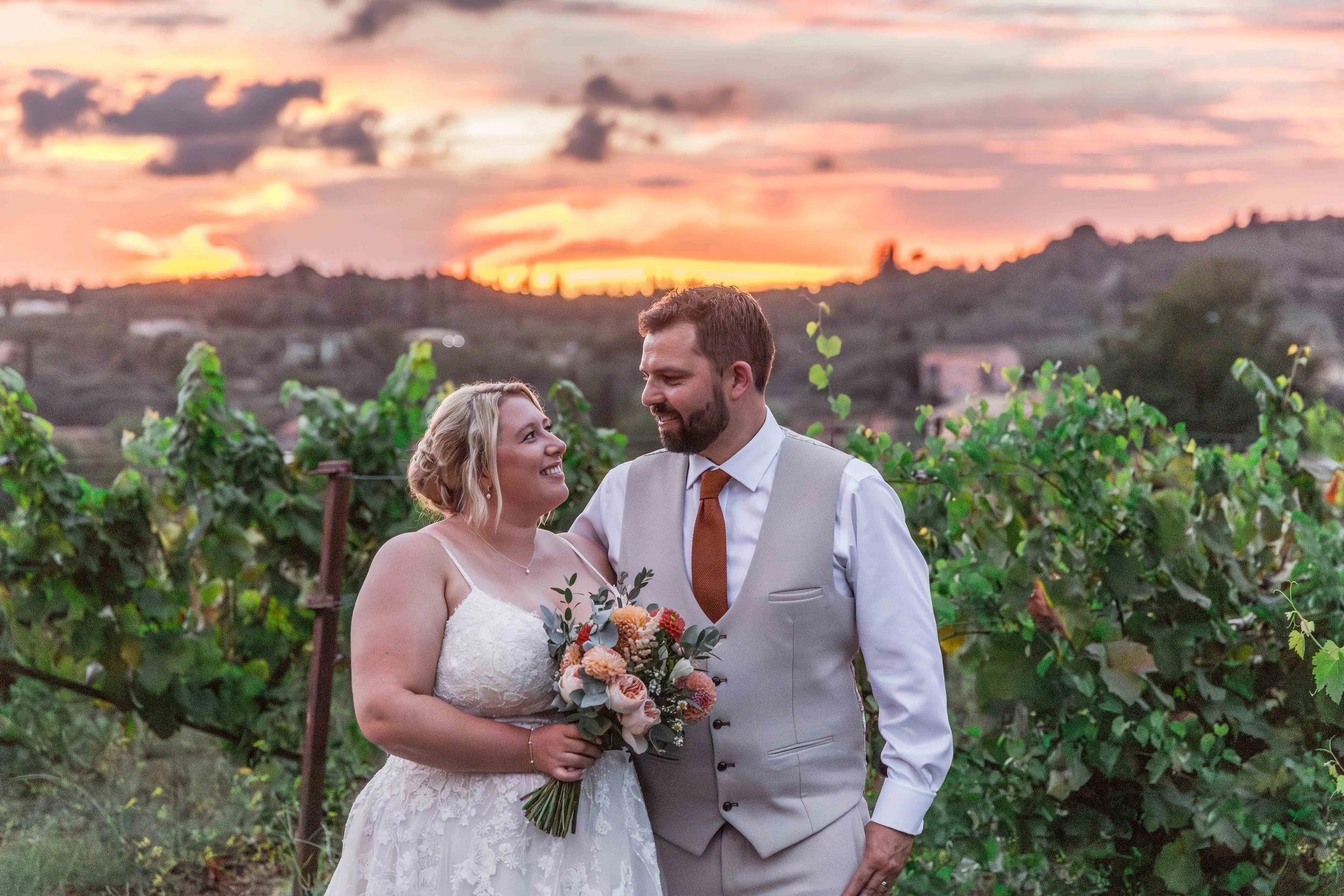 A bride and groom standing close together in a vineyard during sunset, with the bride holding a bouquet of flowers and smiling at the groom.