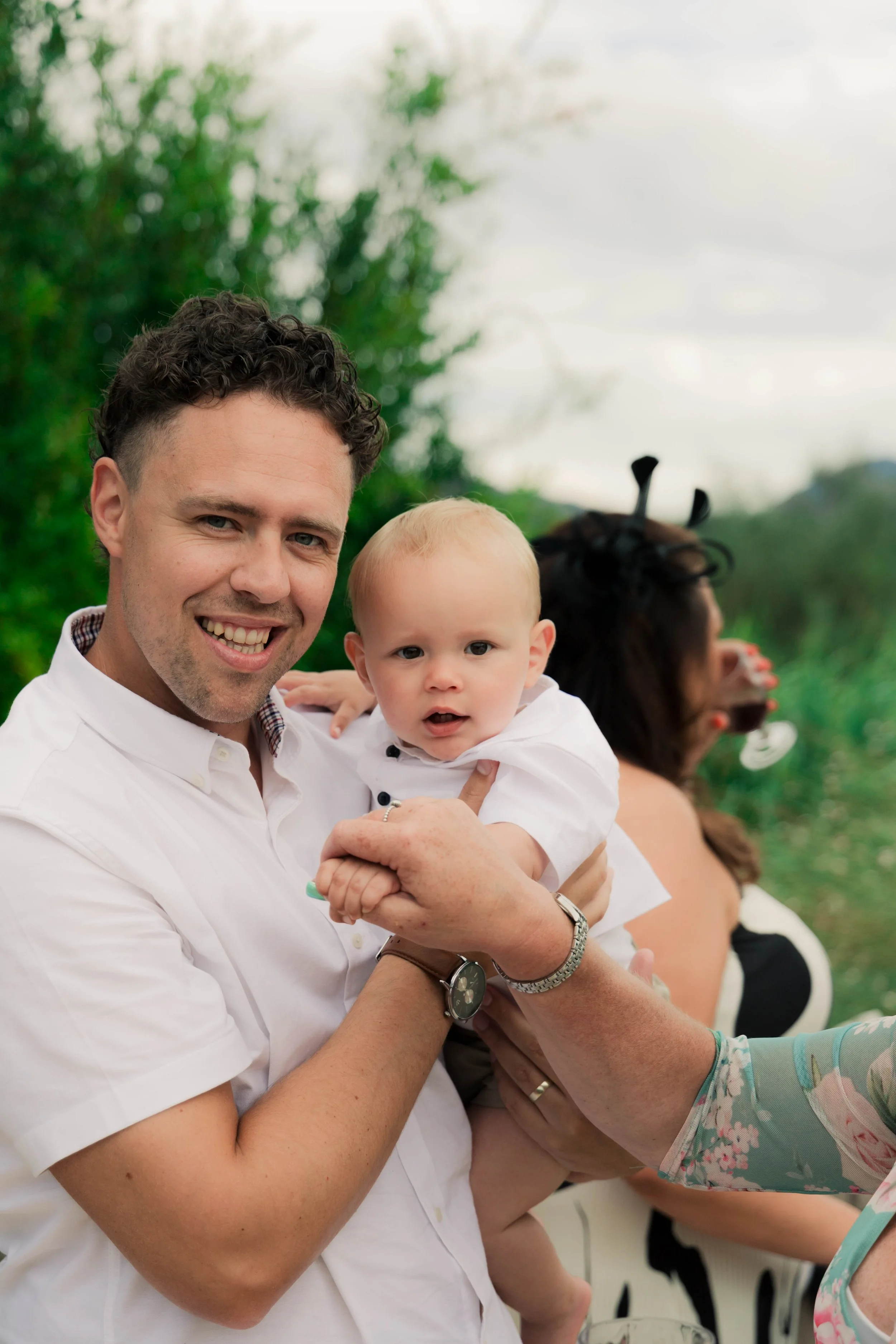 A man smiling while holding a young child with blonde hair and expressive eyes, outdoors with greenery in the background. A woman wearing black bunny ears and drinking a beverage is partially visible in the background.