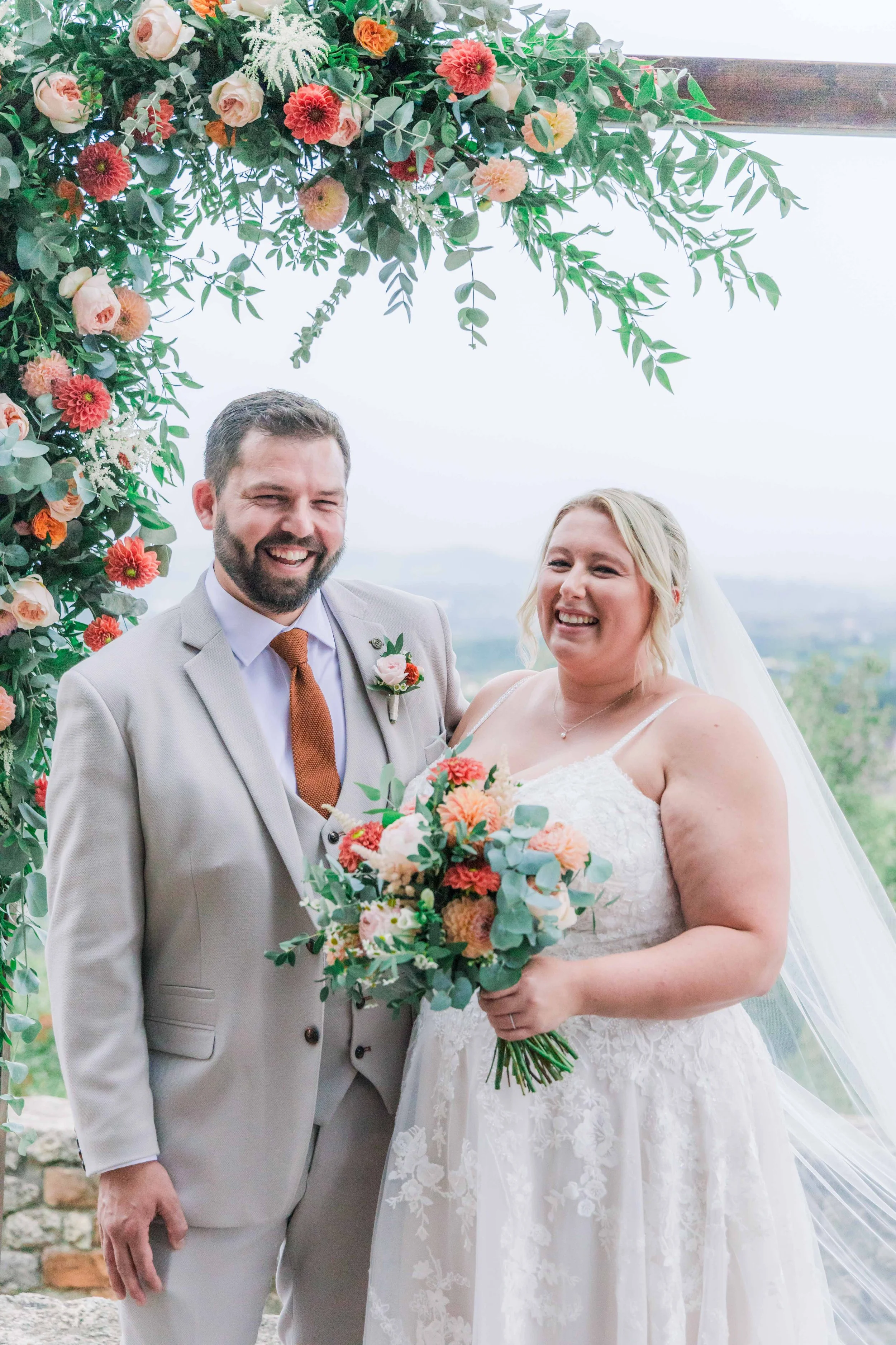 A bride and groom standing under a floral arch during their wedding ceremony, smiling happily. The bride is holding a bouquet of flowers.
