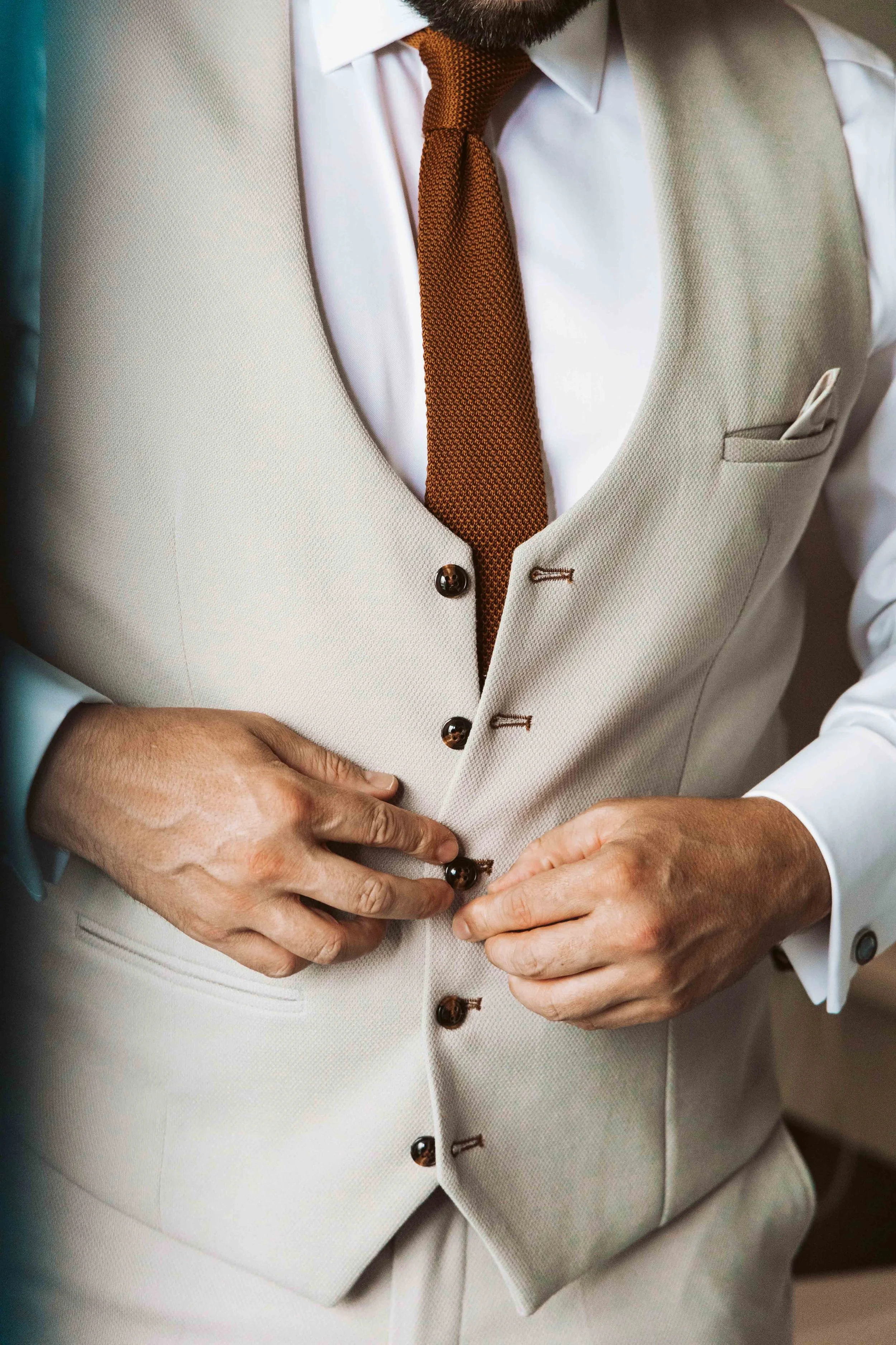 Close-up of a man in a beige suit vest with a white dress shirt and a brown tie, fastening buttons on his vest.