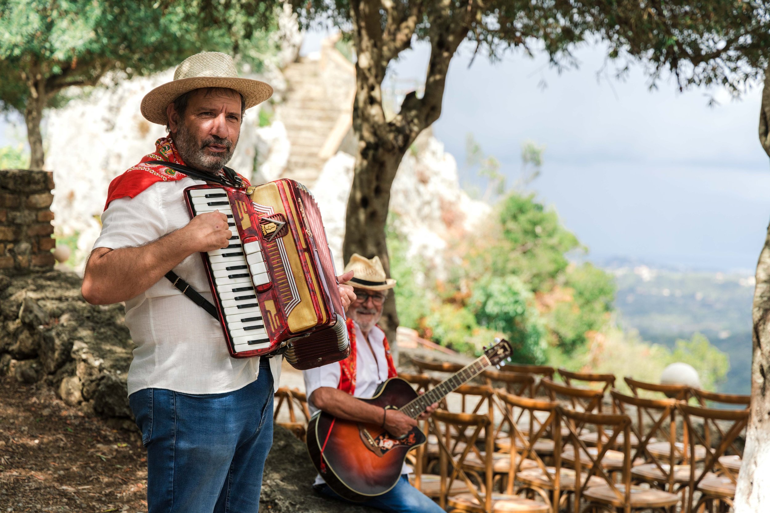 Two men playing musical instruments outdoors during daytime, with a backdrop of trees and landscape. One man wears a straw hat and holds an accordion, while the other has a guitar and wears a fedora-style hat.