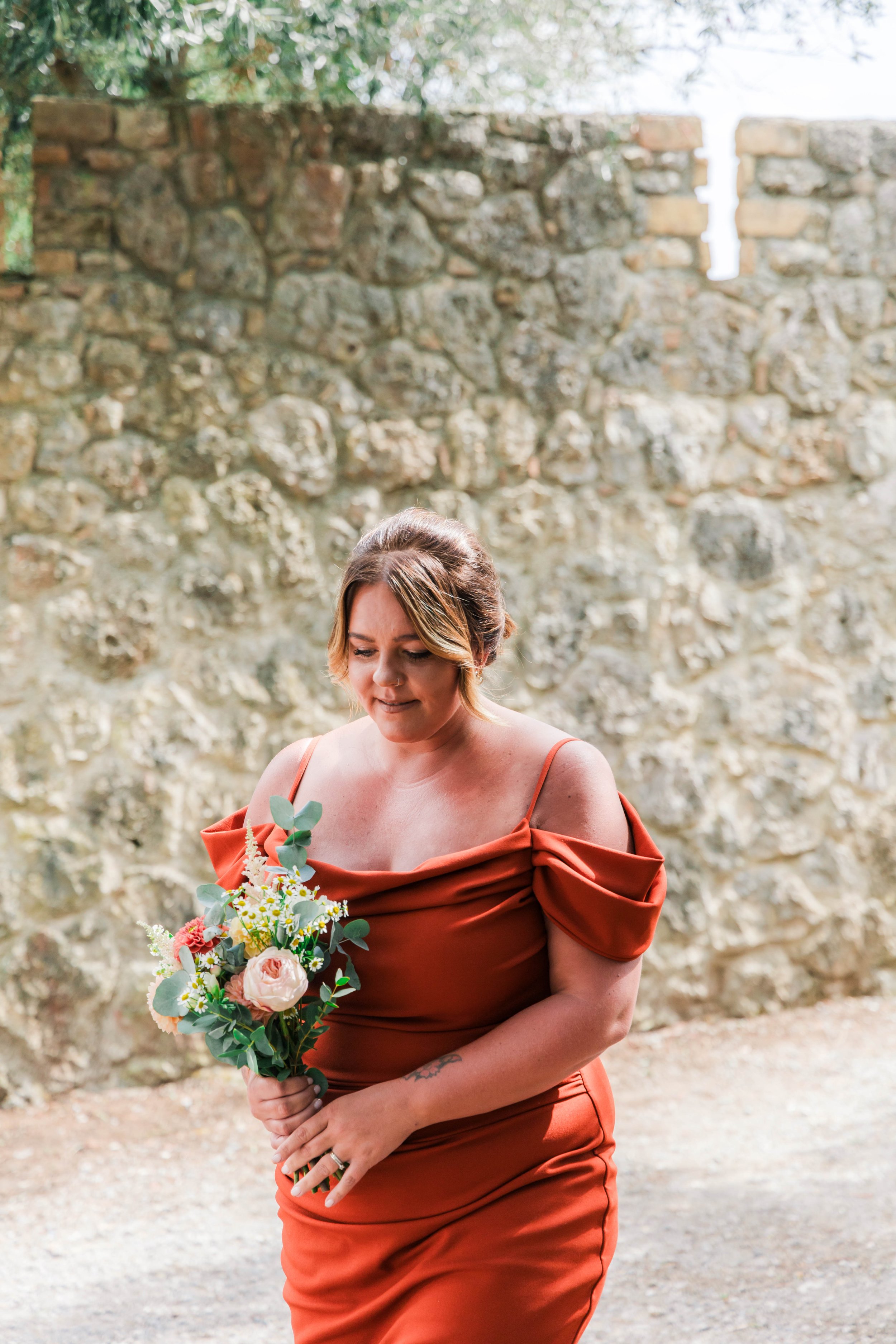 A woman in a rust-colored dress holding a bouquet of pink, white, and green flowers standing outdoors in front of a stone wall.