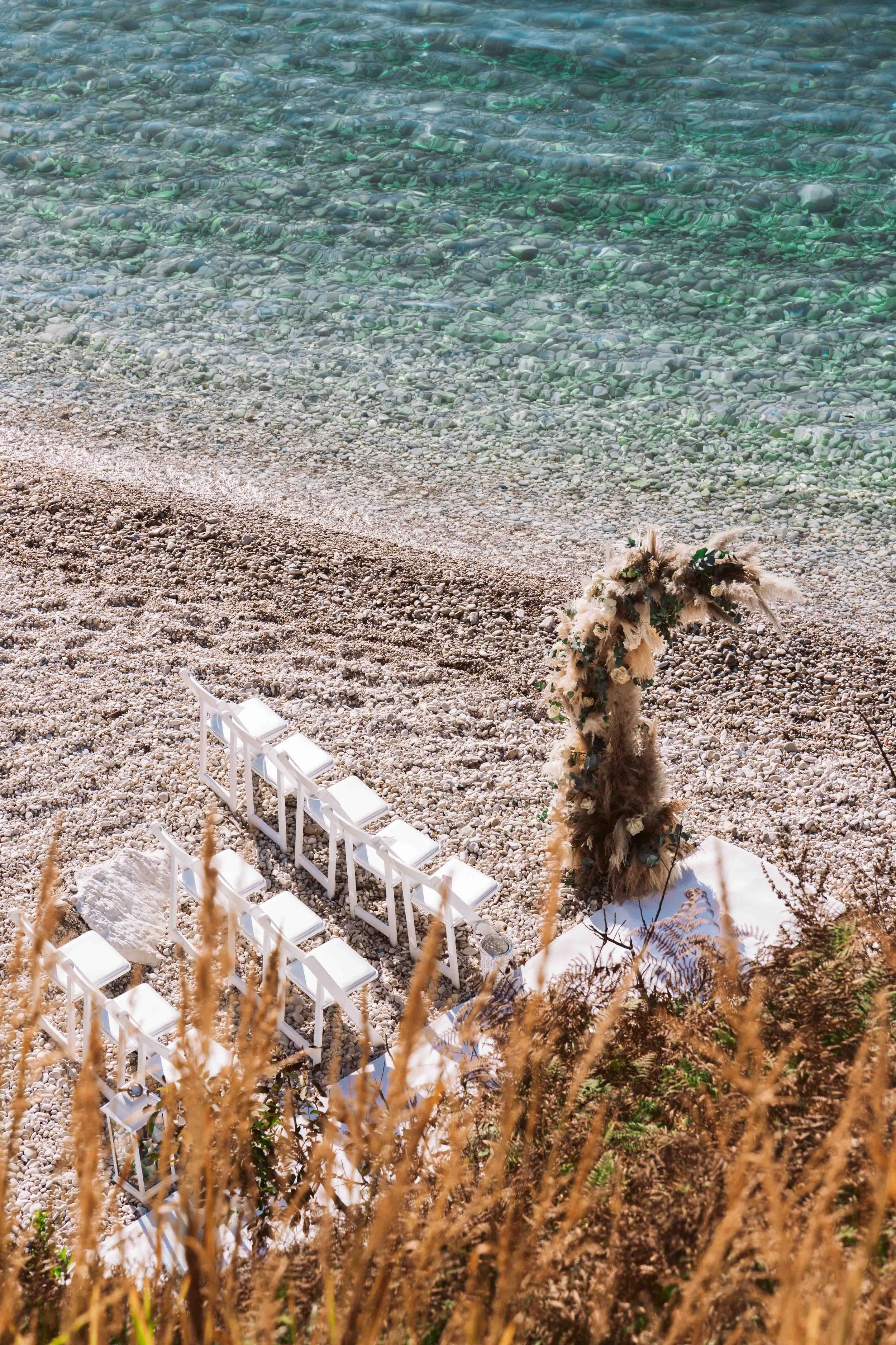Empty white chairs arranged on a pebbled beach near clear green water, with a decorative floral arch and dried grasses in the foreground.