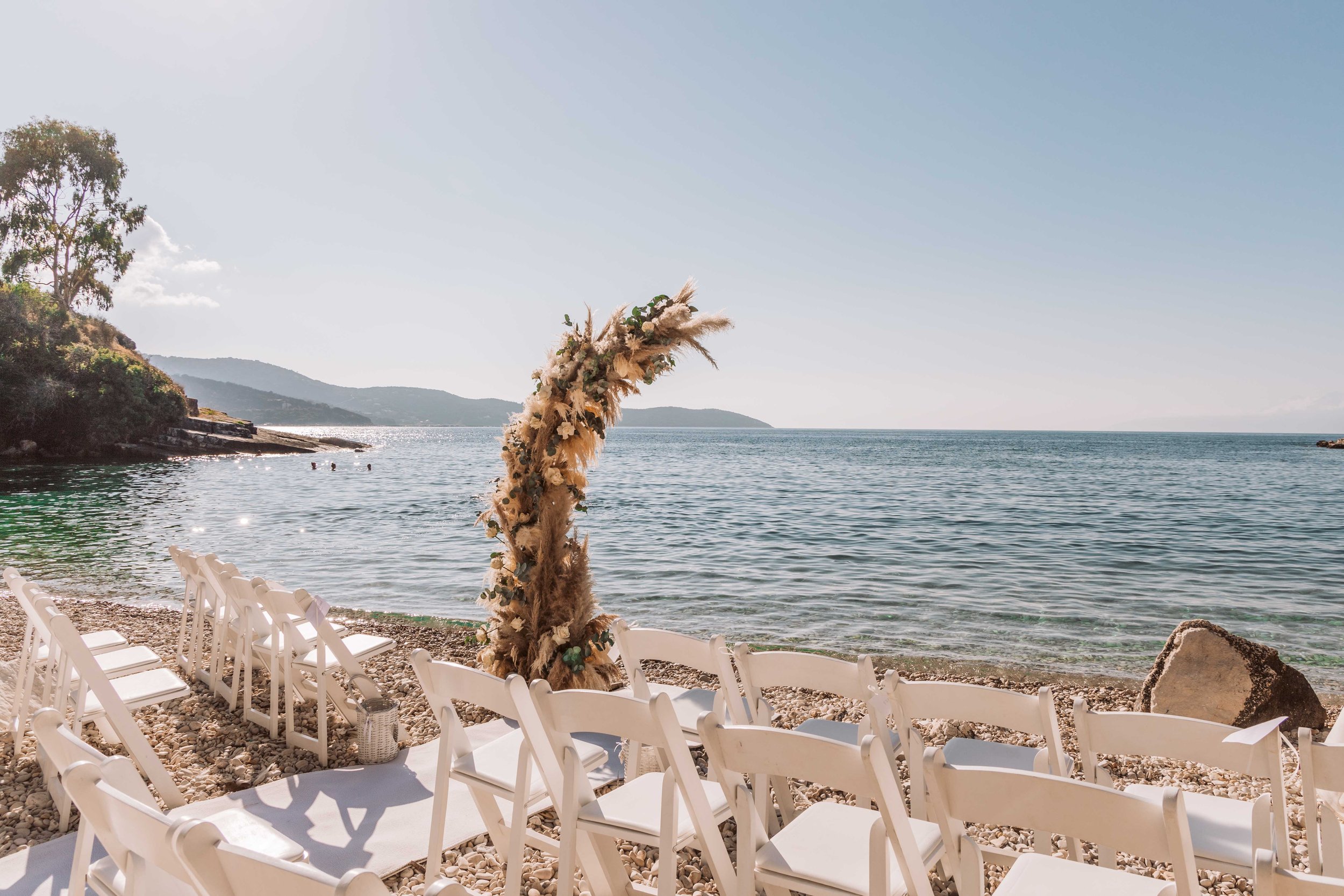 A beach setup with rows of white chairs facing the ocean, and a decorative arch made of dried flowers and greenery, set against a scenic coastal backdrop with calm water, rocky shoreline, and distant hills under clear sky.