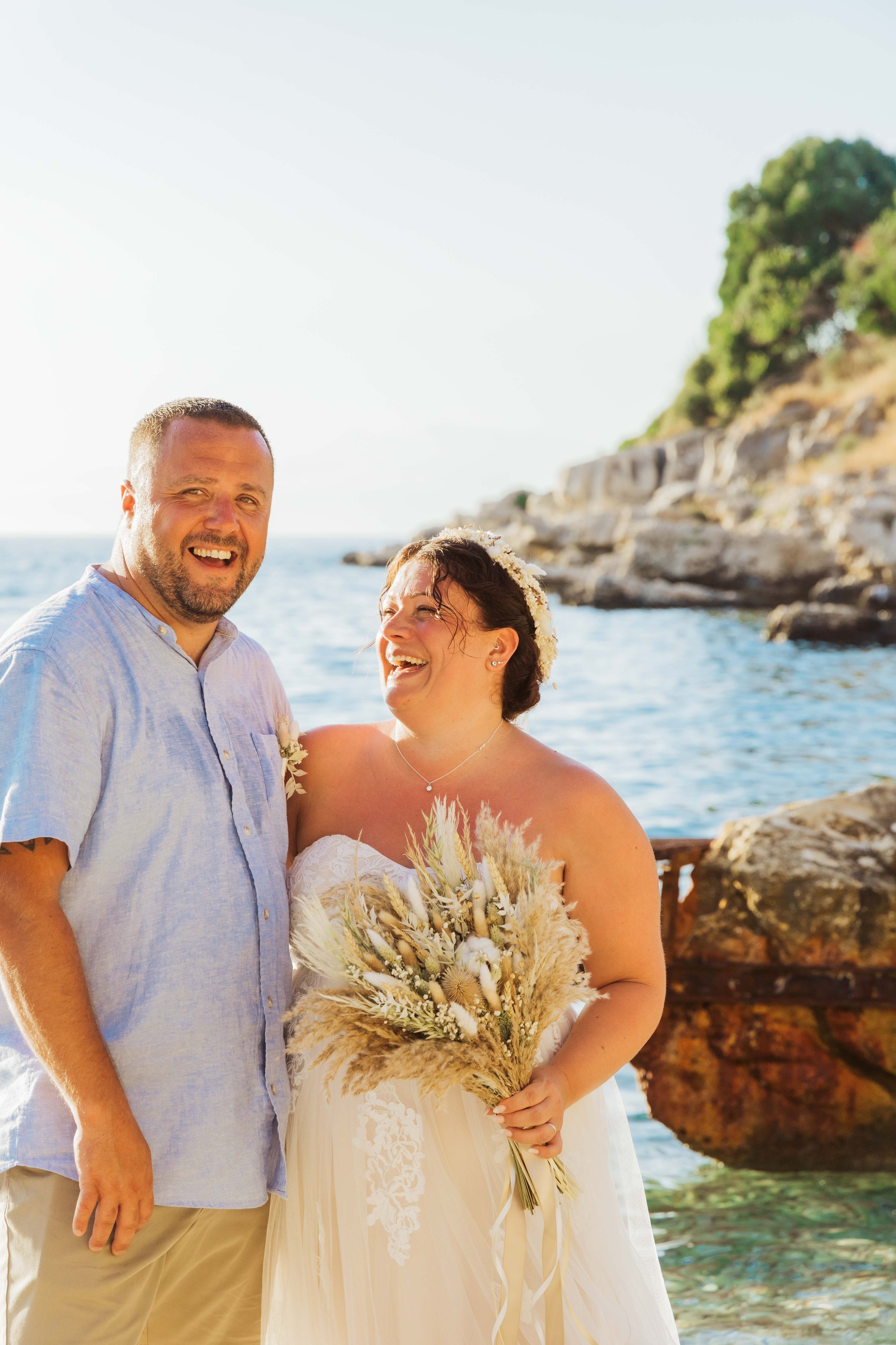 A happy couple at their wedding, standing by the water with rocky cliffs and greenery in the background, the bride holding a bouquet of dried flowers.