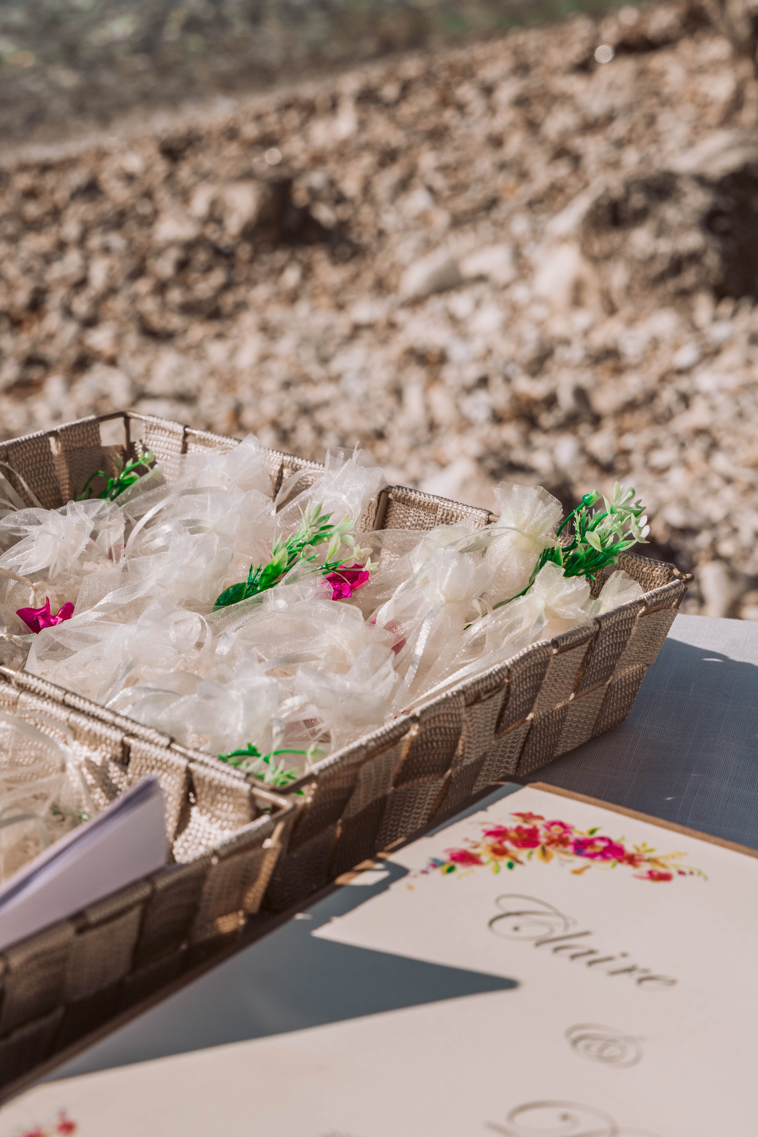 A woven basket containing small gift bags wrapped in clear cellophane with green and pink decorative accents. Part of a white card with colorful flowers and cursive writing is visible in the corner, set on a table outdoors with a rocky ground in the 
