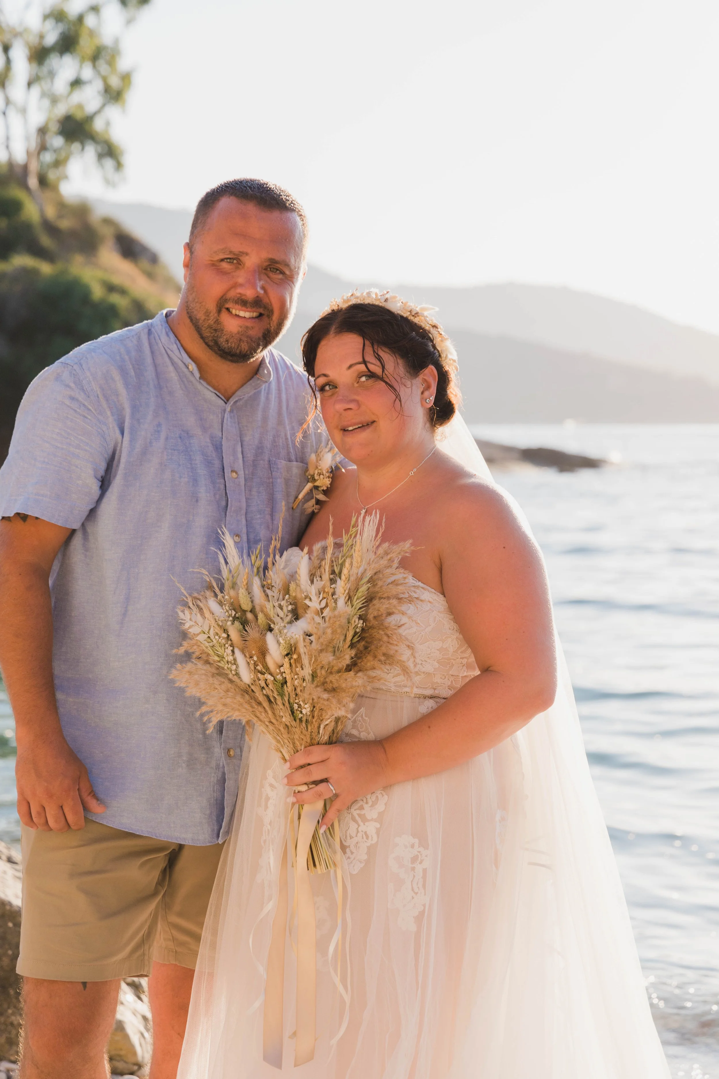A couple dressed in wedding attire standing by the beach at sunset, with the woman holding a bouquet of dried flowers.