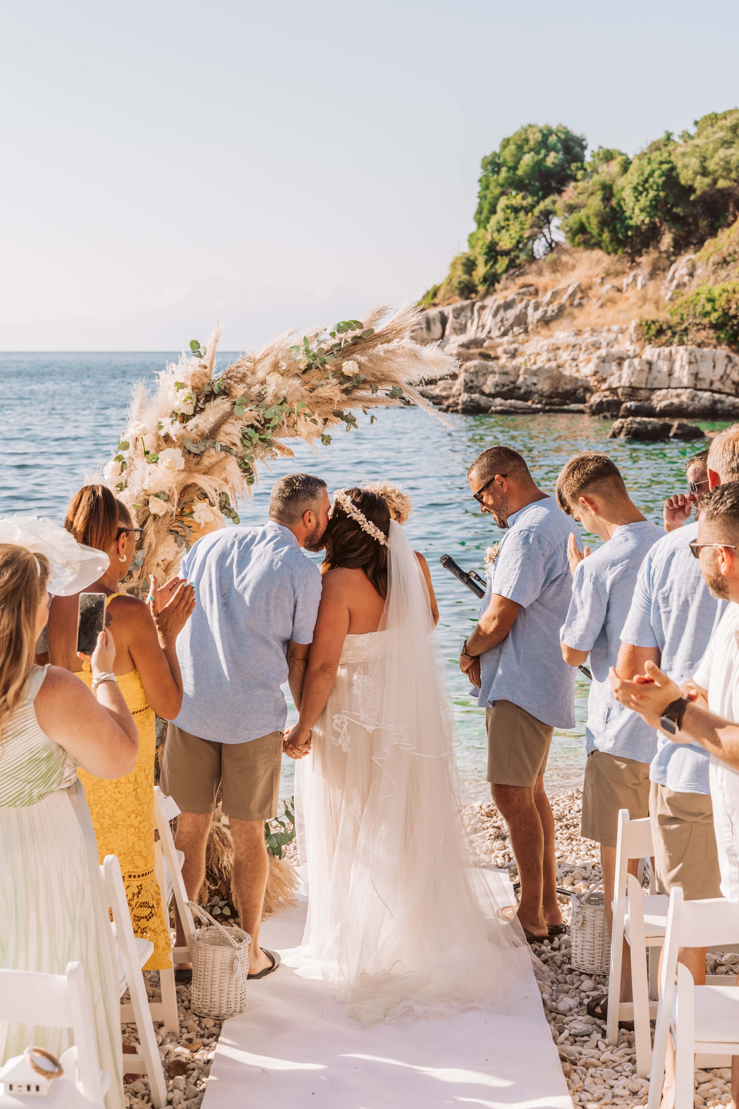 A wedding ceremony by the seaside with a bride and groom kissing under a floral arch surrounded by friends and family.