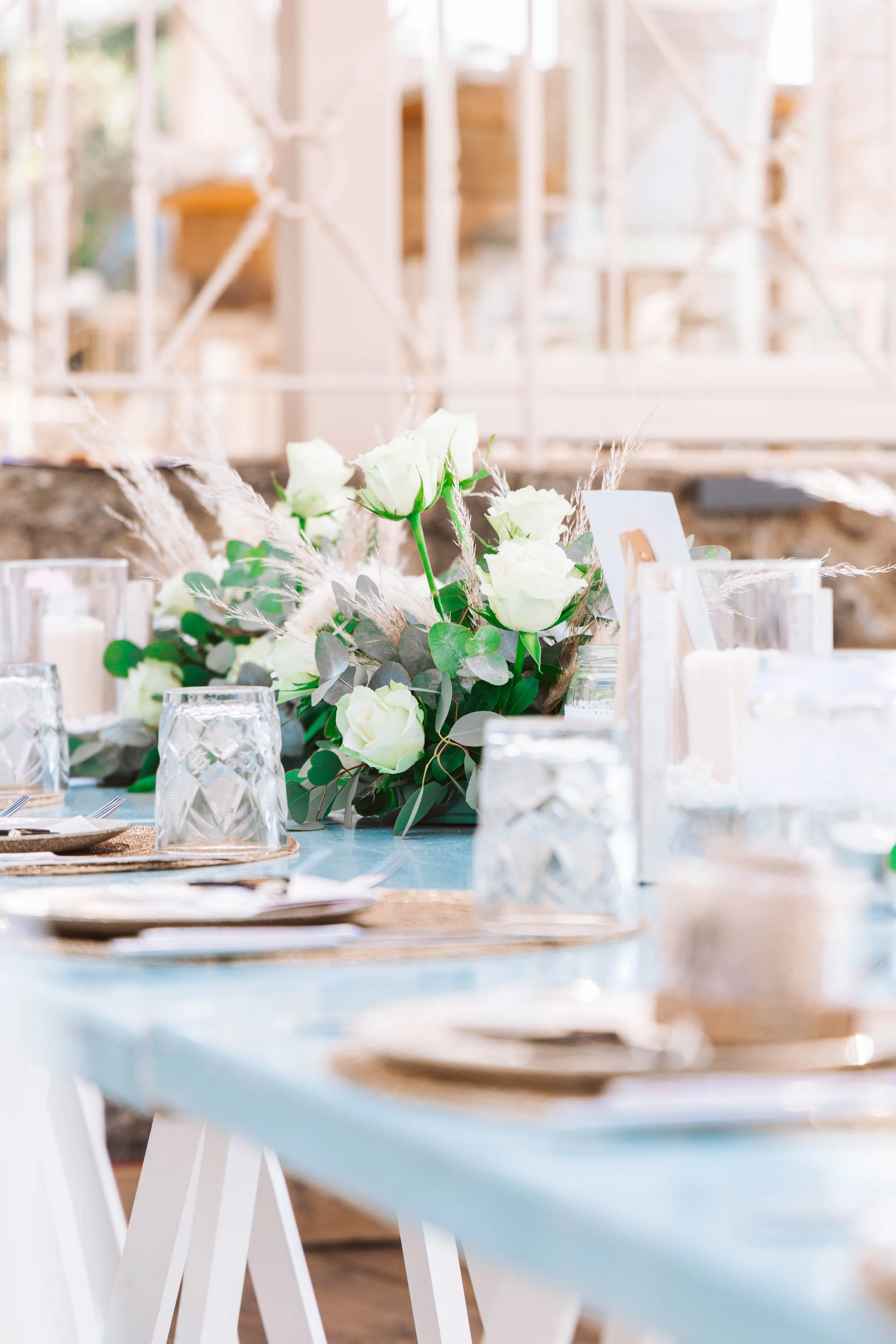 A table decorated with a floral centerpiece of white roses and greenery, set with plates, glasses, and cutlery in a bright, modern setting.