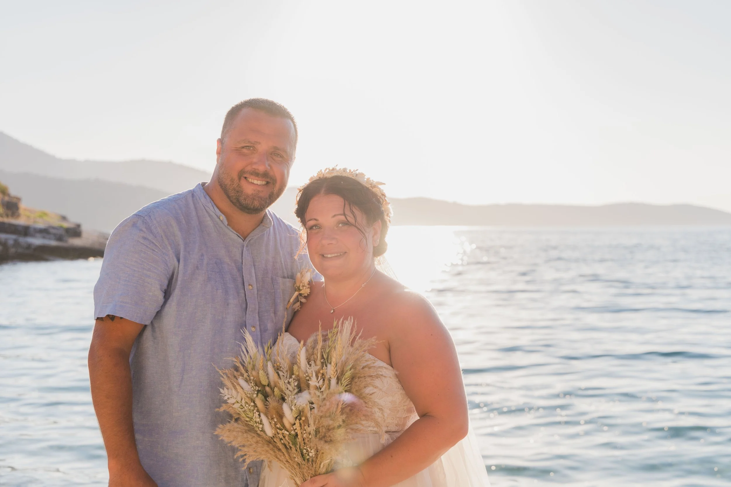 A happy couple standing together by the water during sunset, with mountains in the background. The woman is holding a bouquet of dried flowers and wearing a strapless dress, while the man is dressed in a light blue, short-sleeved shirt.