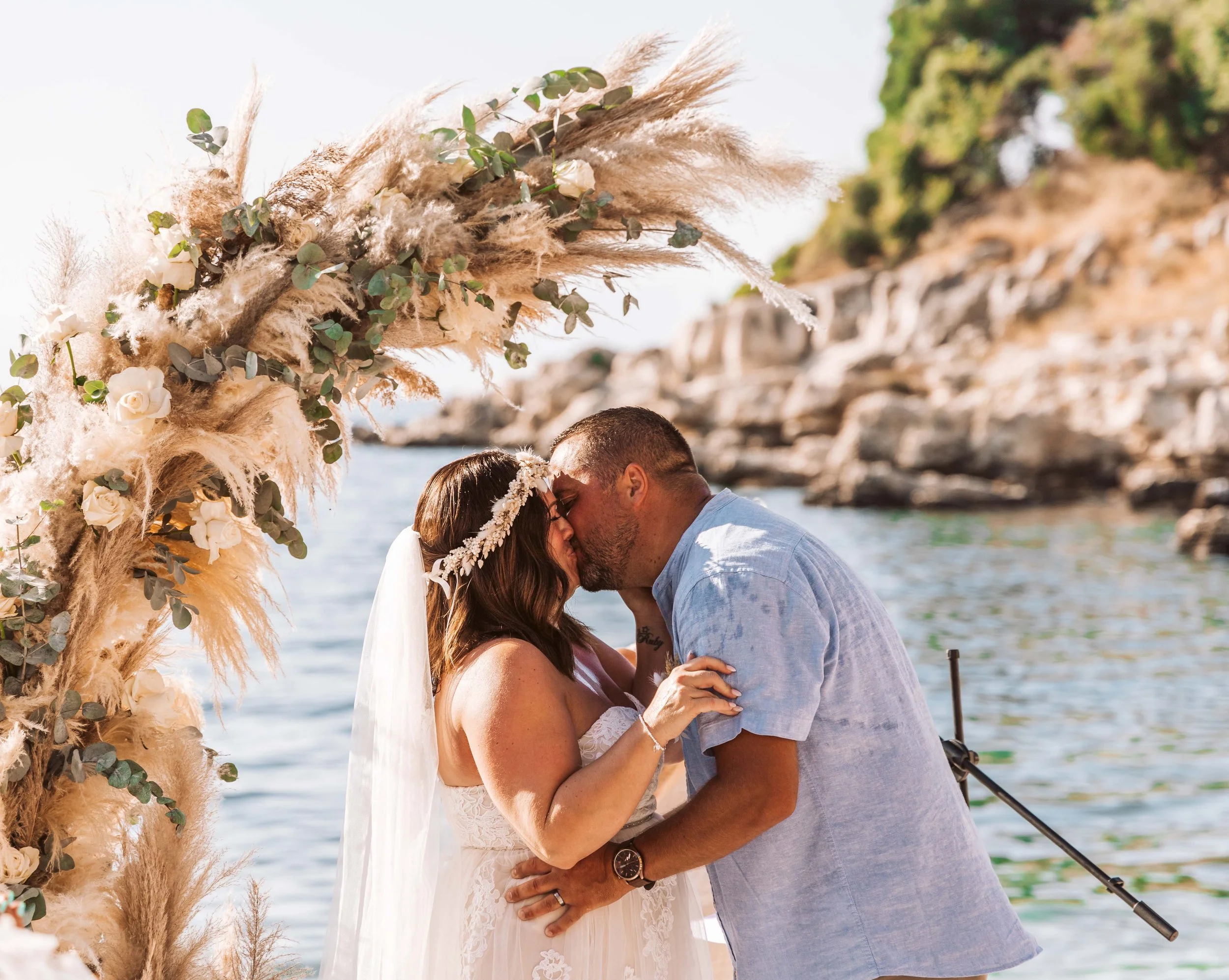 A couple sharing a kiss during their wedding ceremony by the water, with a decorative floral arch and rocky shoreline in the background.