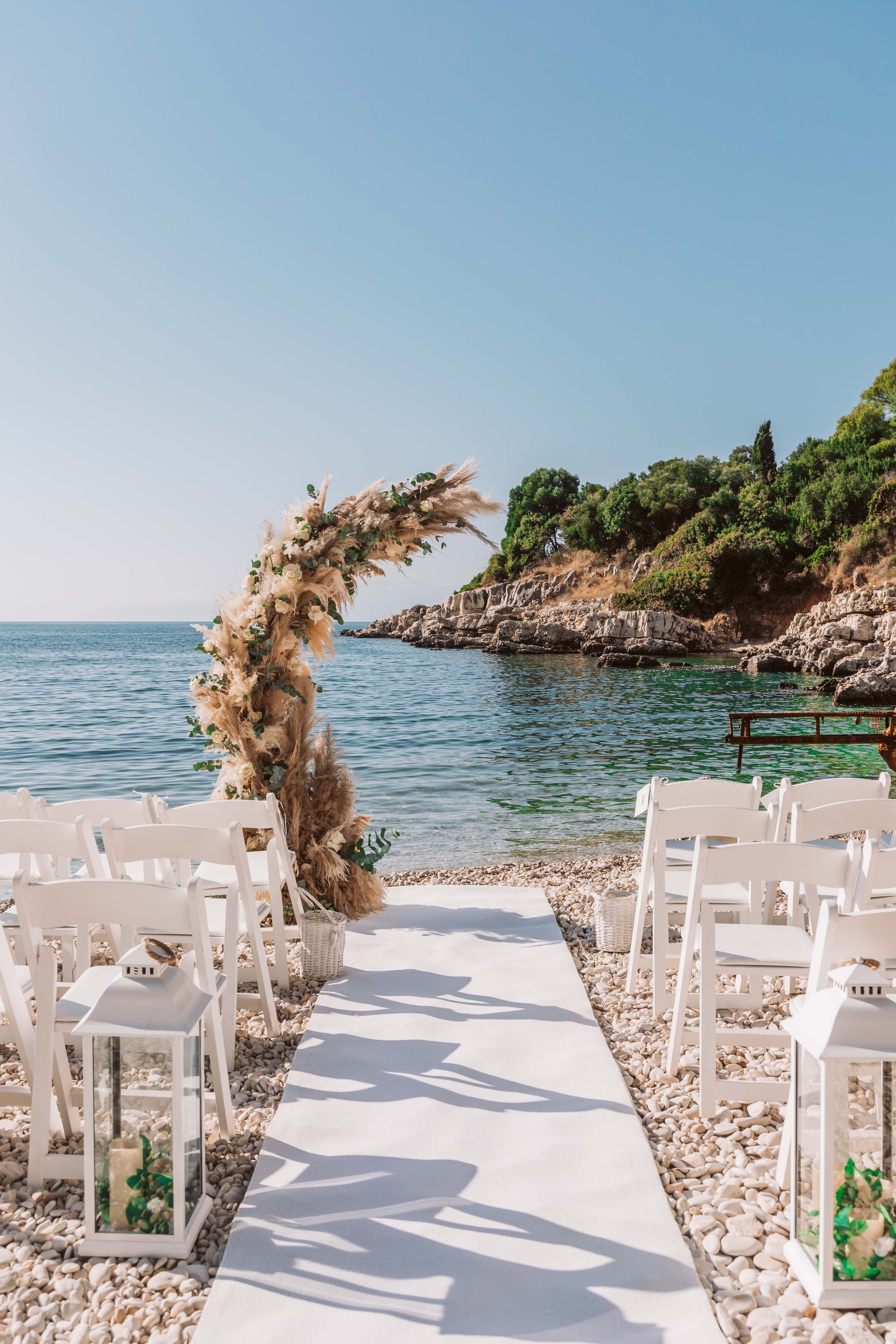 Beach wedding setup with white chairs, a fabric aisle, a floral arch, and lanterns, overlooking the ocean and rocky shoreline under a clear sky.