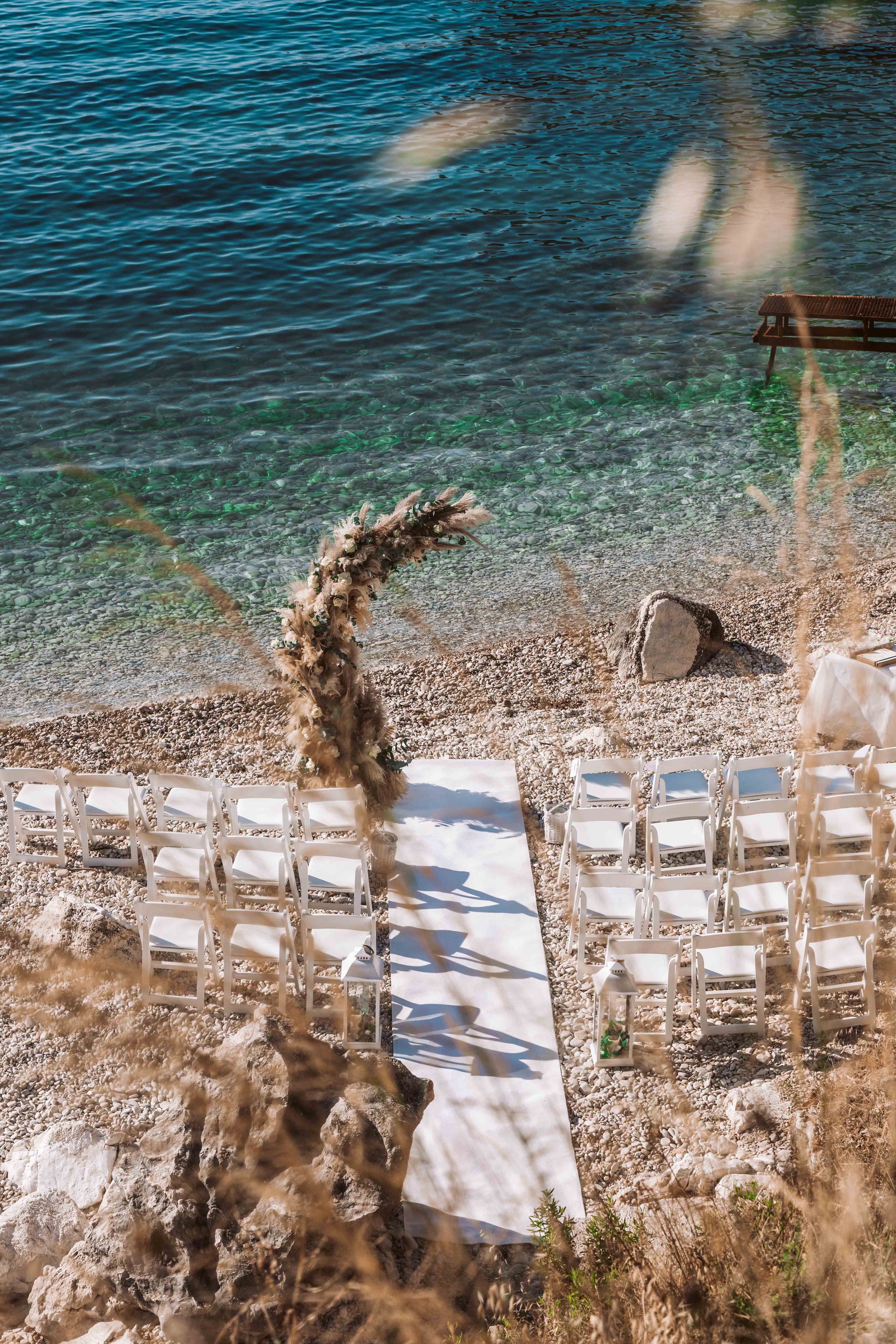 Empty white chairs arranged along a white aisle runner on a rocky beach near turquoise water; floral arch at the select altar area, gentle waves, grassy plants, and rocks in the background.