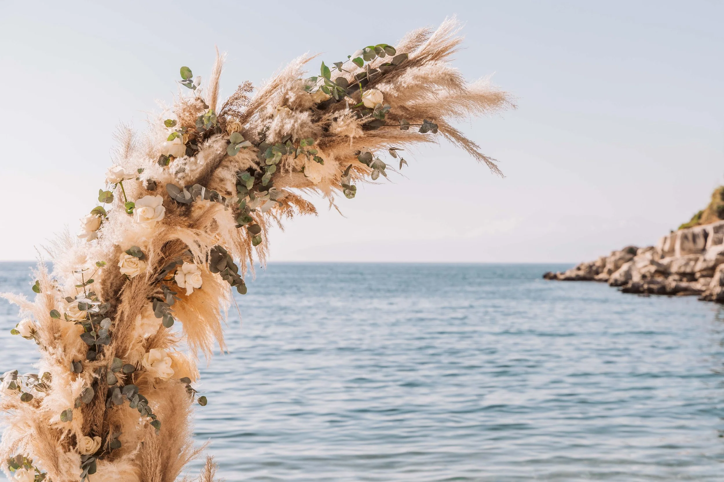 A decorative floral arrangement with pampas grass and white roses along a coastline with water and rocky cliffs in the background.