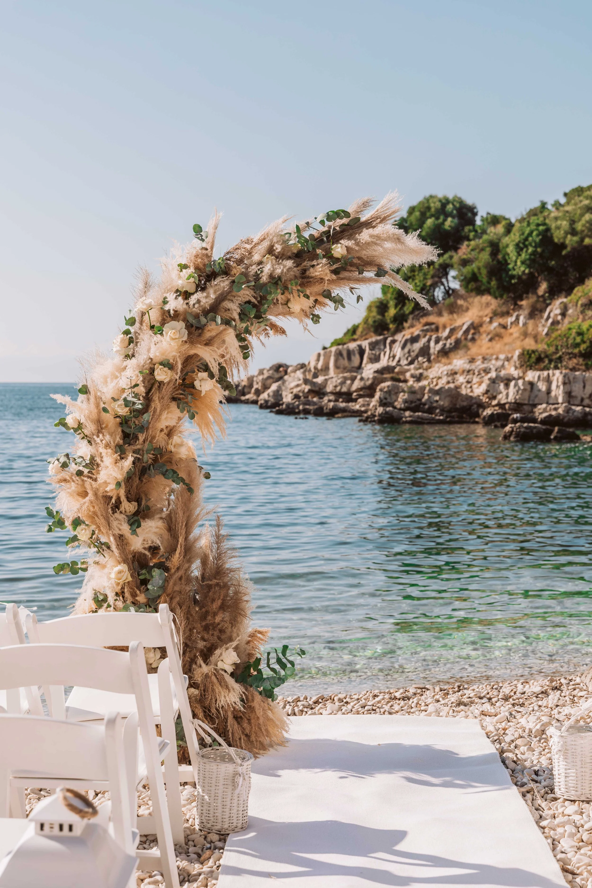 Beachside wedding arch decorated with pampas grass, white roses, and greenery, with white chairs and a white aisle runner on pebbled shore, overlooking water and rocky cliffs under a clear sky.