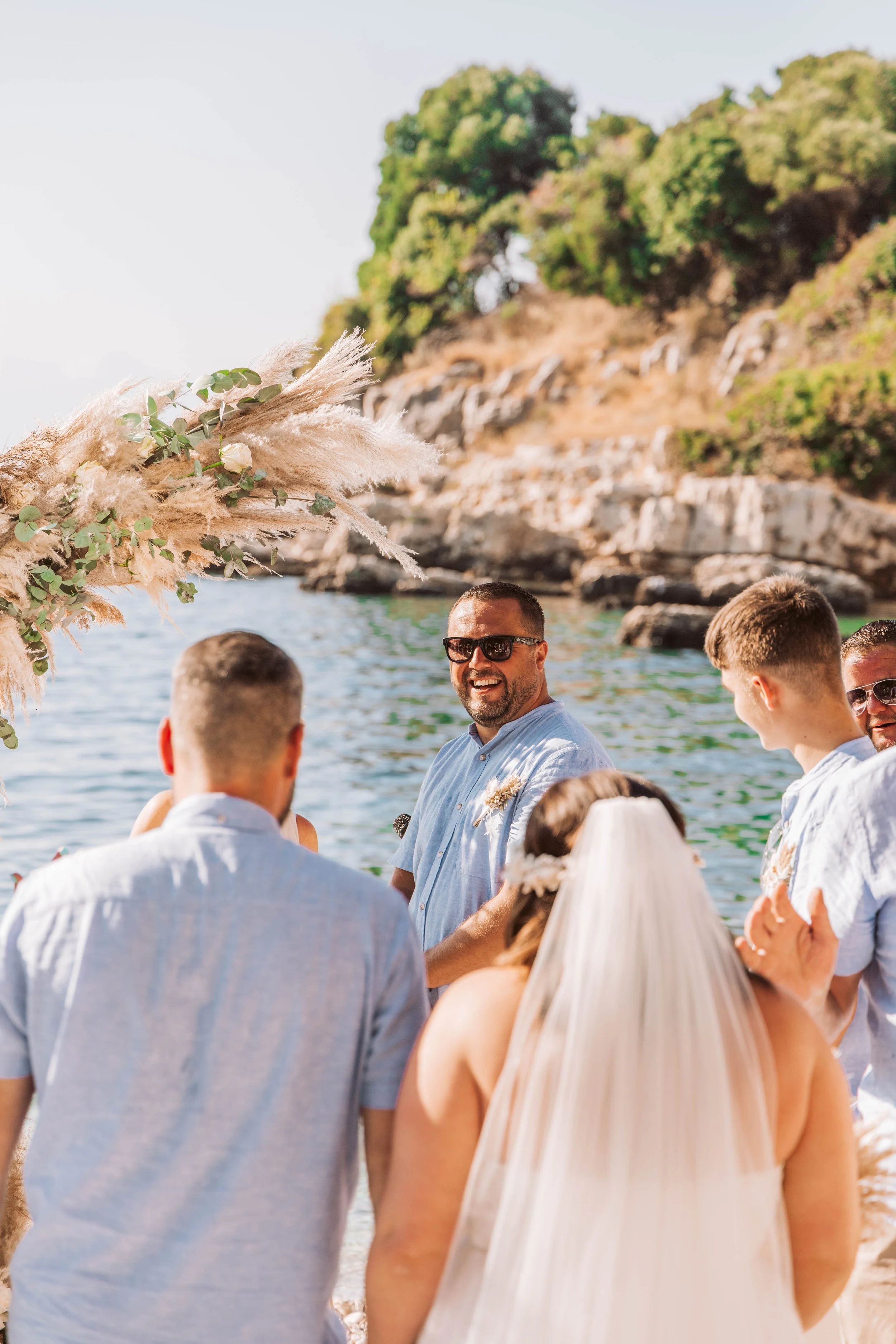 A group of people at a wedding on a beach, with a man smiling and wearing sunglasses, surrounded by friends or family, with rocky cliffs and greenery in the background.