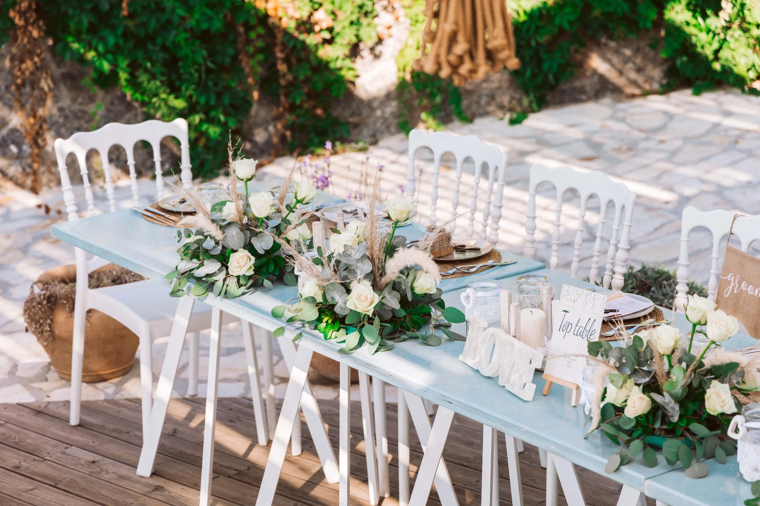 A decorated outdoor table with a floral centerpiece of white roses and green foliage, surrounded by white chairs and set for a celebration, possibly a wedding, with signs indicating the 'Top table' and 'Groom'.