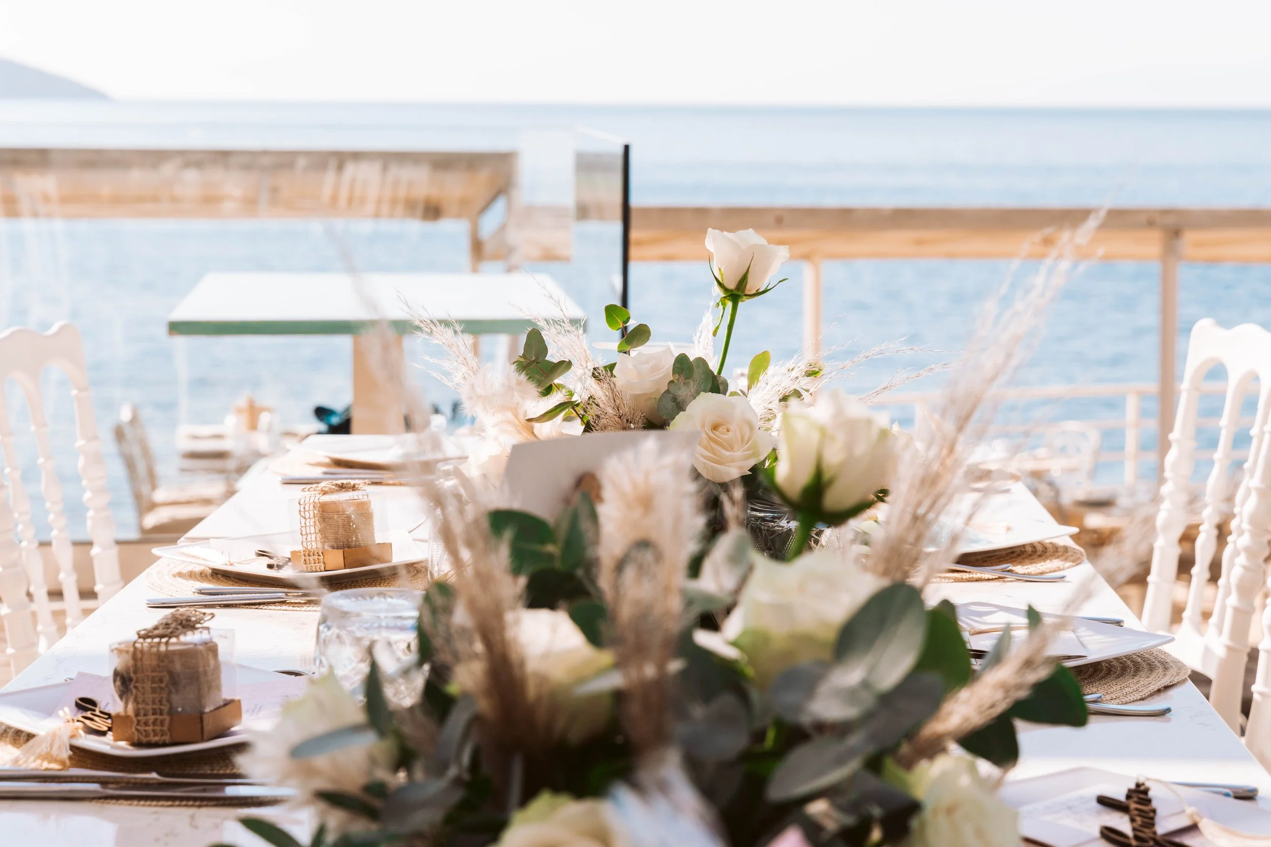 A decorated outdoor dining table set near a body of water with a floral centerpiece featuring white roses and greenery, and a wooden deck in the background.