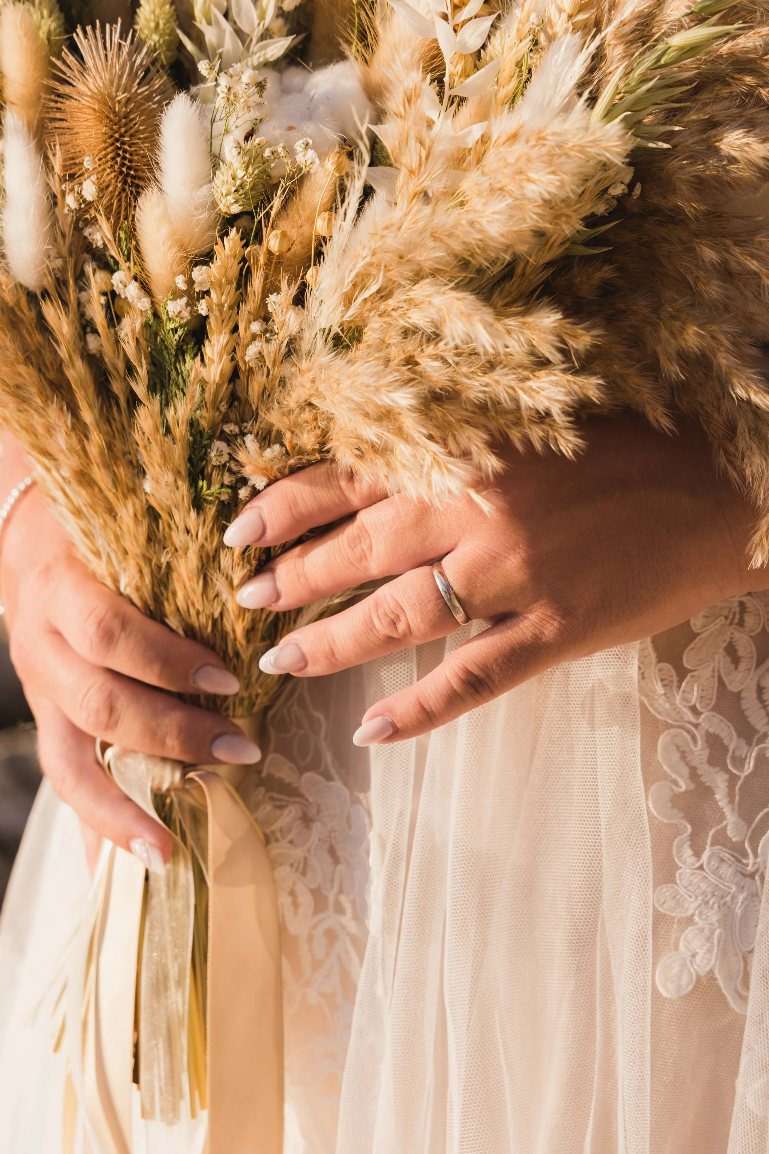 Close-up of a bride's hands holding a bouquet of dried flowers, including grasses and foliage, with lace wedding dress visible.