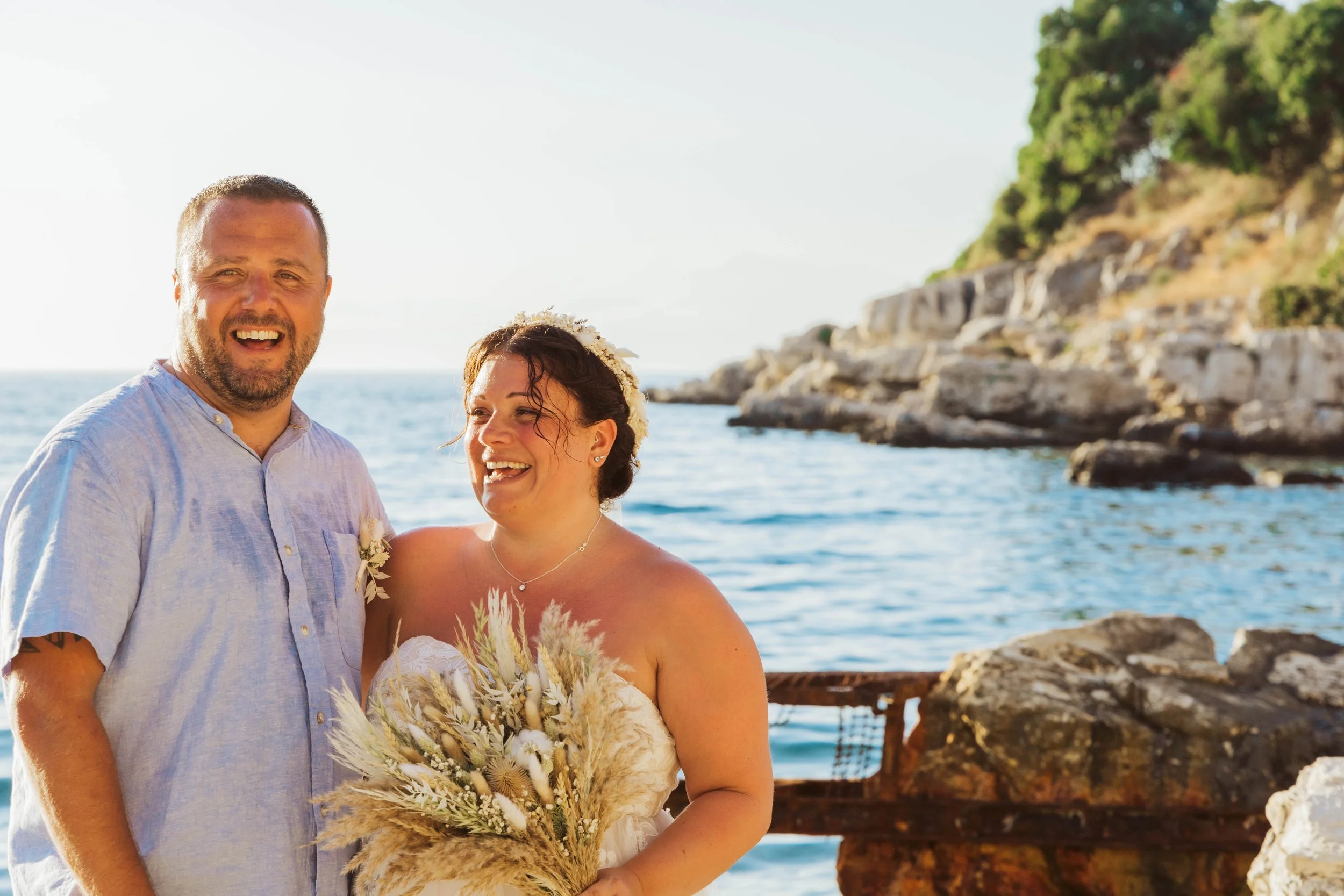 A happy bride and groom standing by the water, smiling, with rocky cliffs and trees in the background, during a wedding photoshoot.