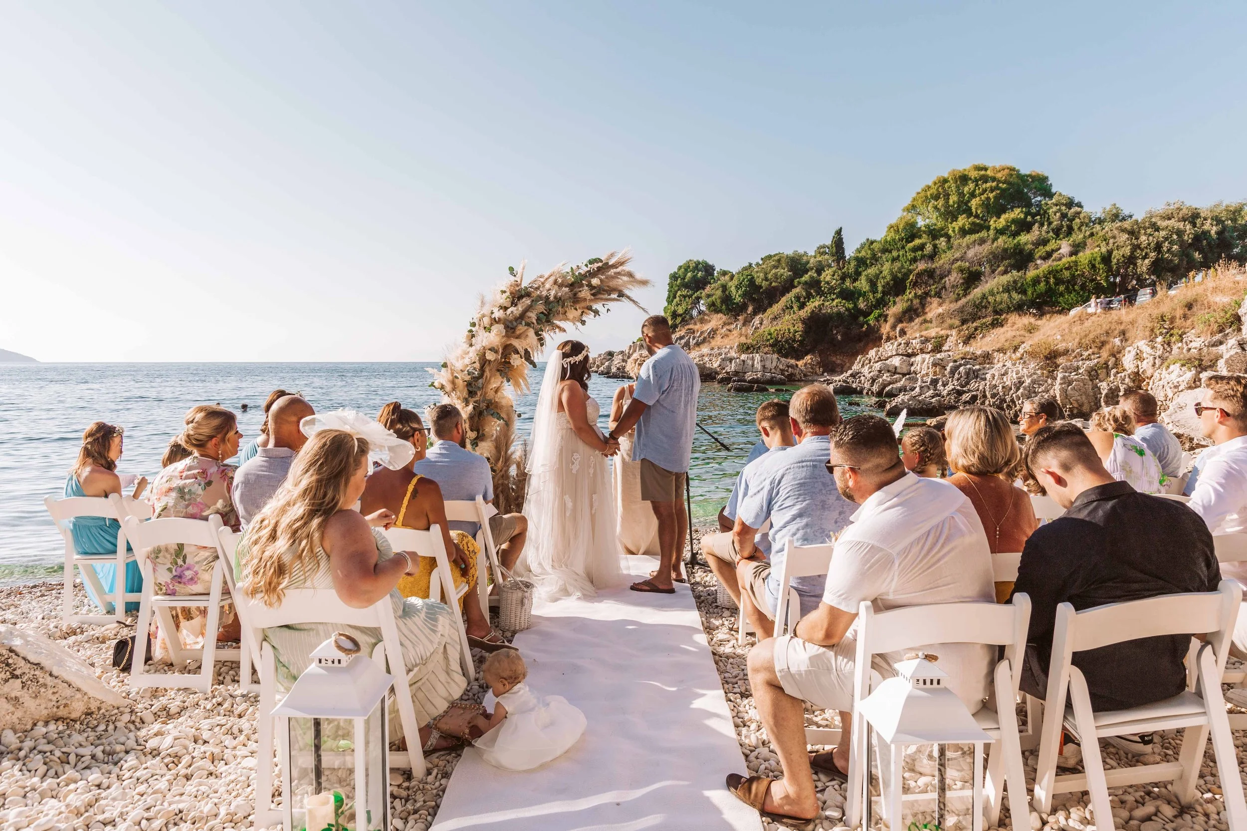 A wedding ceremony taking place on a beach with an ocean and rocky hillside in the background. The bride and groom are holding hands under a floral arch, surrounded by seated guests on white chairs.