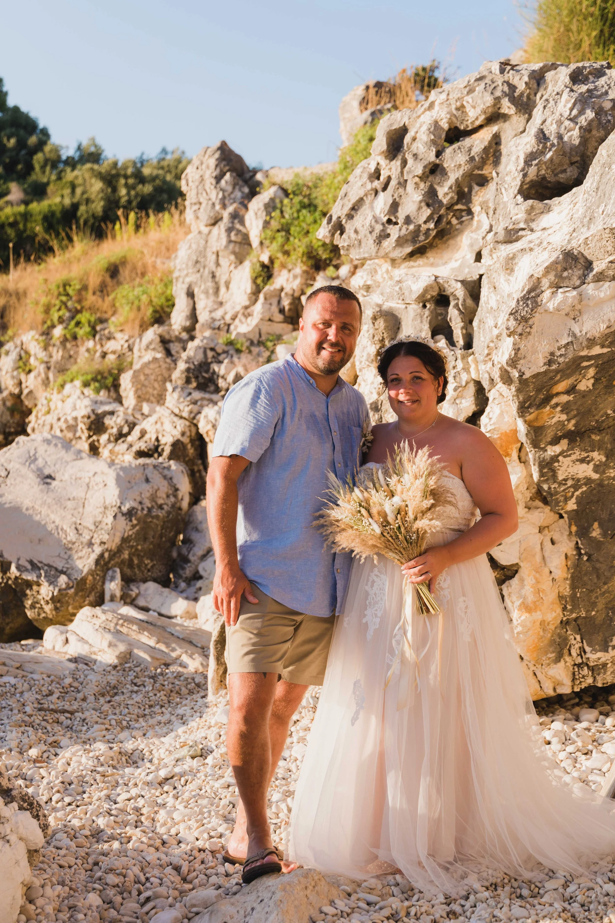 A couple dressed in wedding attire standing on a rocky beach with large rocks and greenery in the background during sunset.