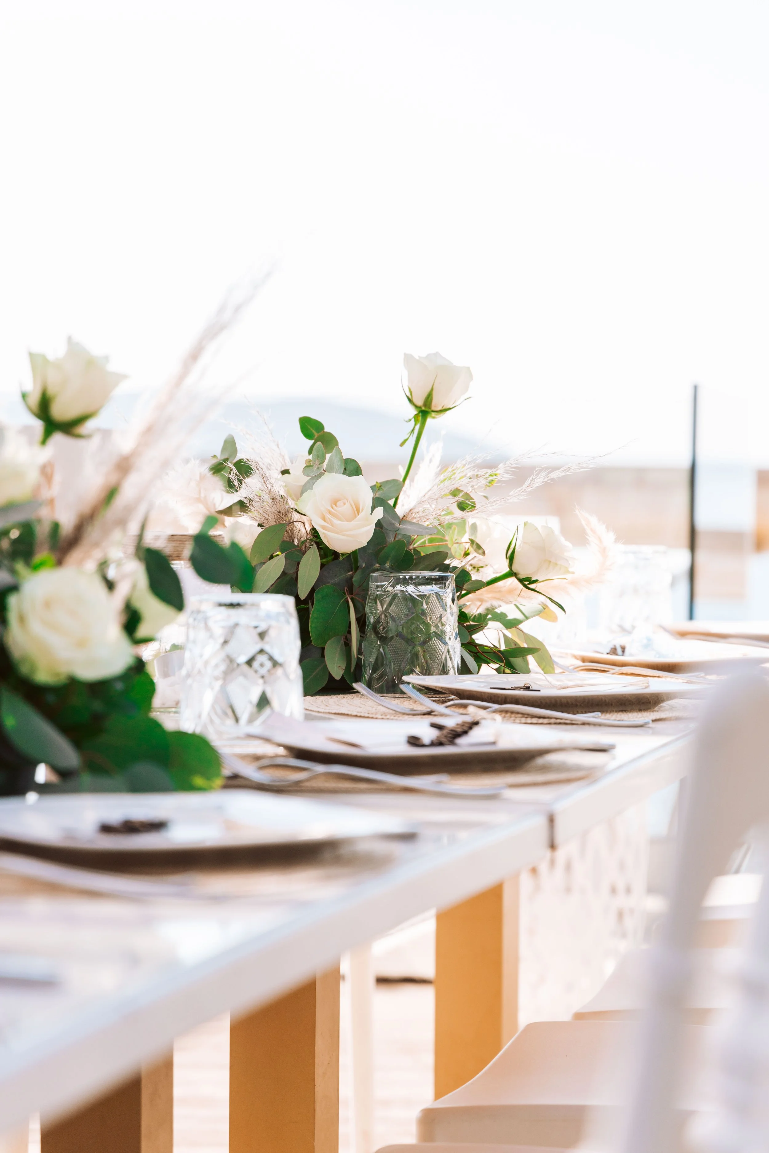 Elegant table setting with white roses, greenery, and decorative glassware in a bright, possibly outdoor, setting.