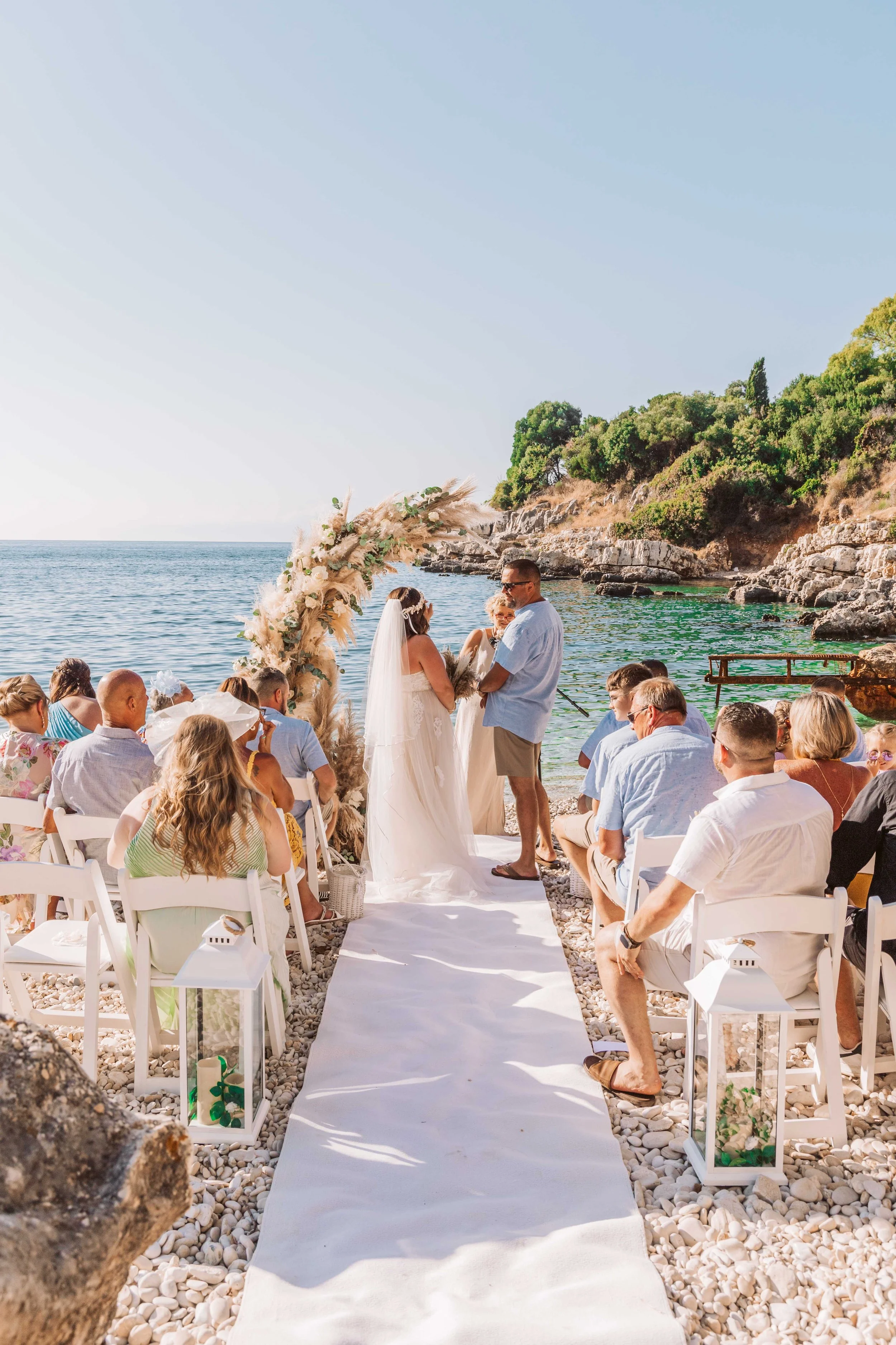 A beach wedding ceremony with guests seated on both sides of a white aisle, a couple exchanging vows under a floral arch, near the ocean with rocks and greenery in the background, on a sunny day.