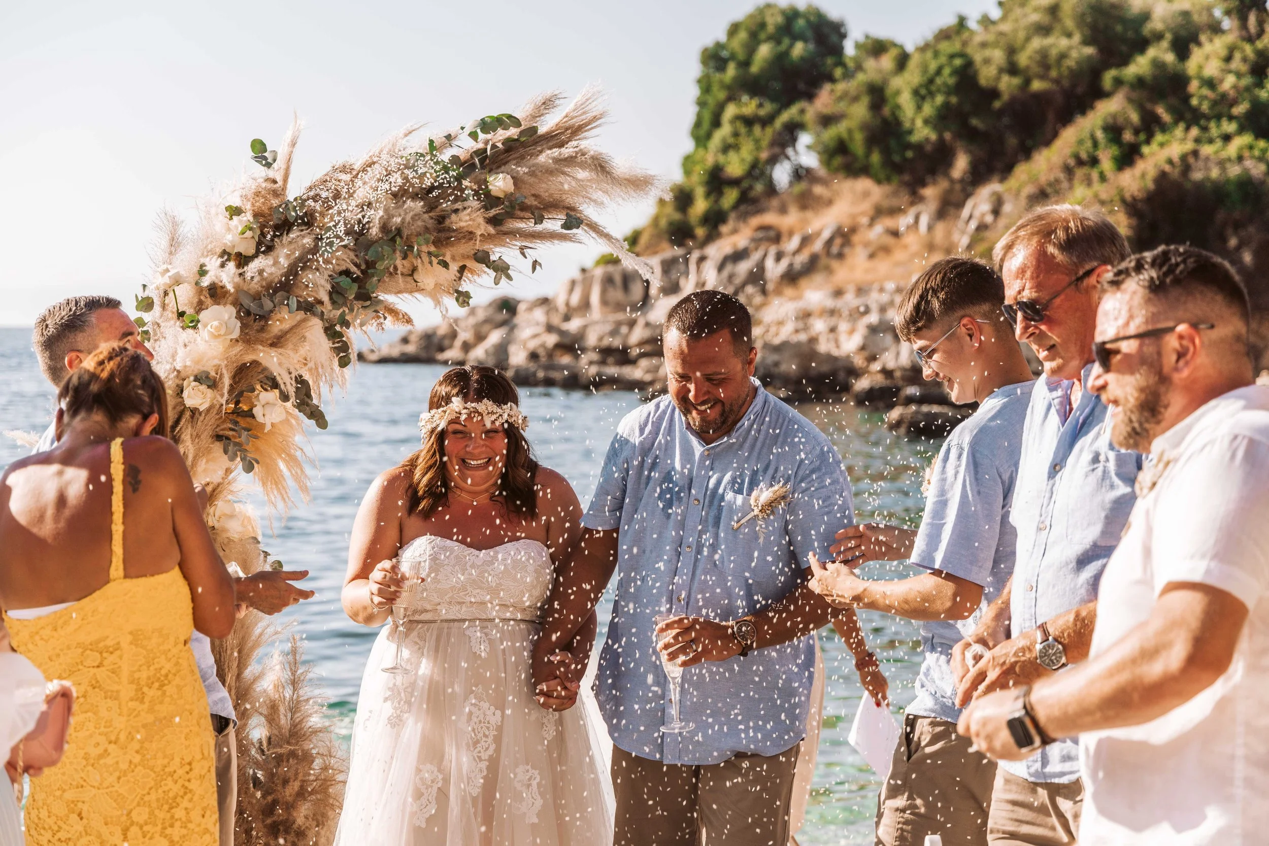 A couple getting married on a beach during daytime, surrounded by friends and family celebrating with confetti and smiles, with rocky hills and the ocean in the background.