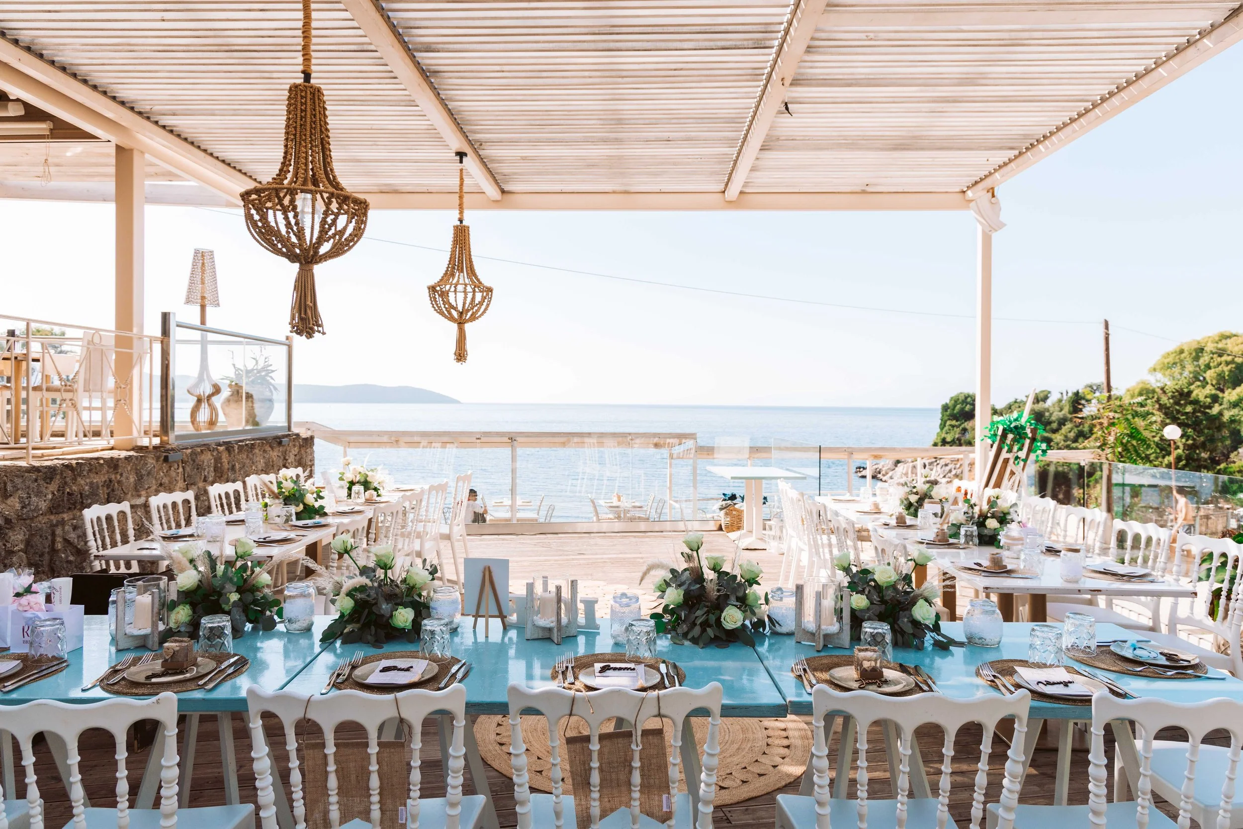 Seafront outdoor dining area decorated for a celebration with floral arrangements, candles, and table settings, overlooking the ocean under a covered patio.