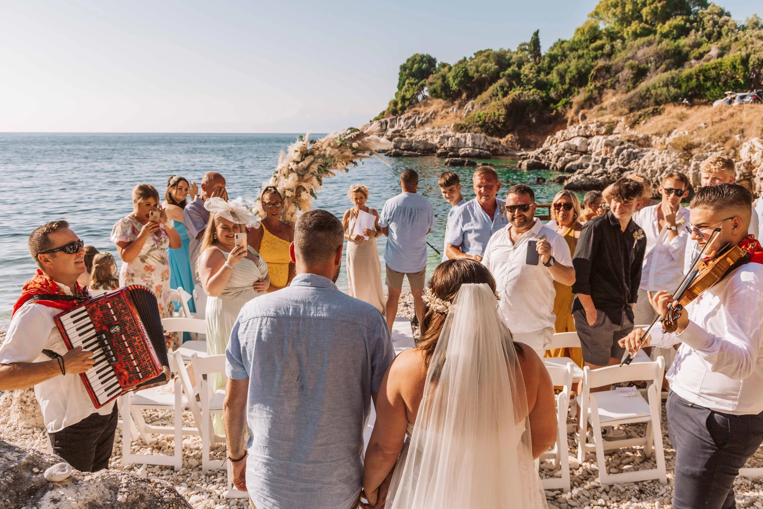 A wedding ceremony taking place on a beach with guests and musicians, overlooking the ocean and rocky cliffs.