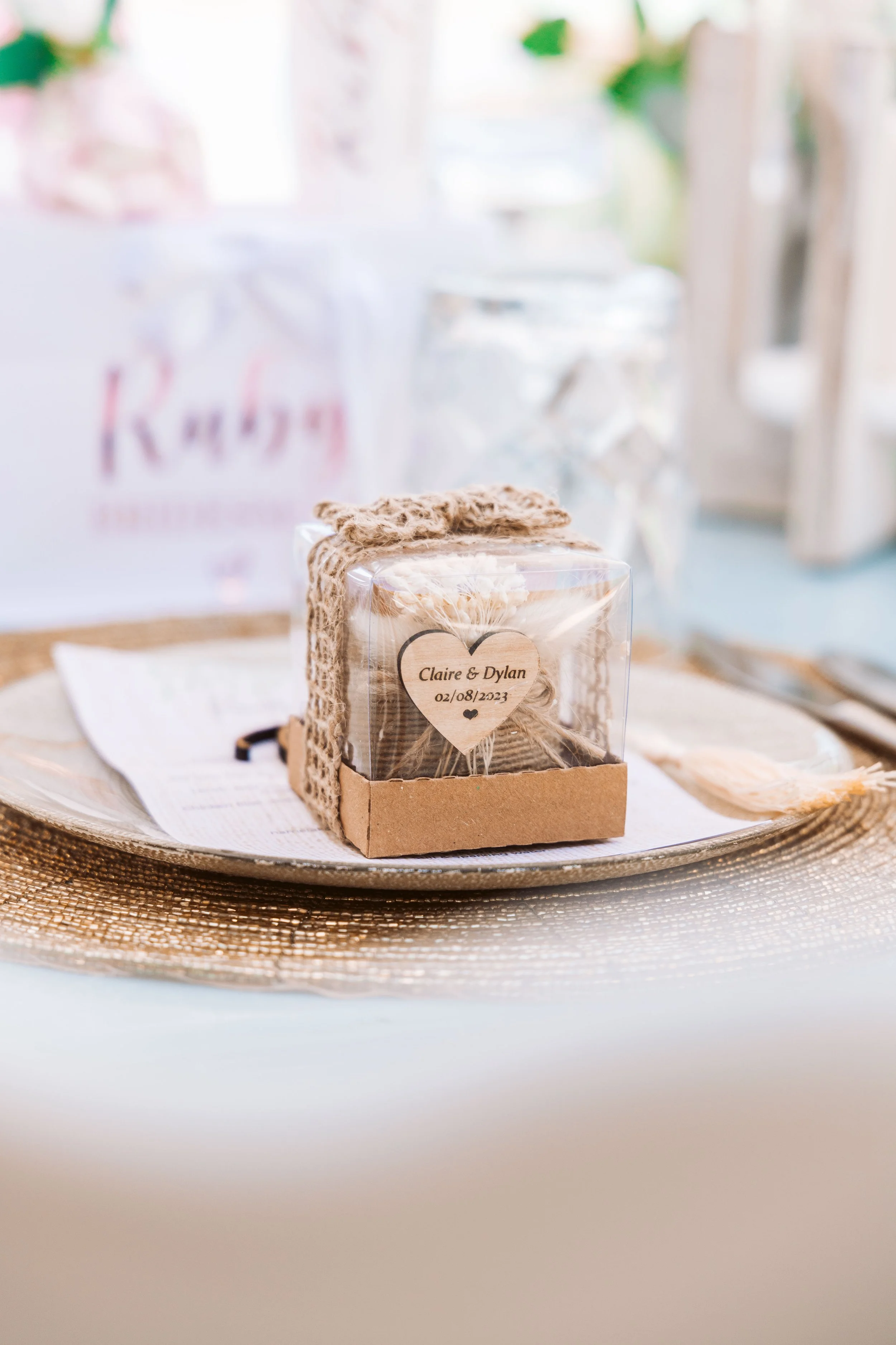 A small clear box decorated with lace ribbon contains a cookie or small gift, placed on a decorative gold placemat on a table set for a special occasion, with a wedding or event details on the label inside the box.