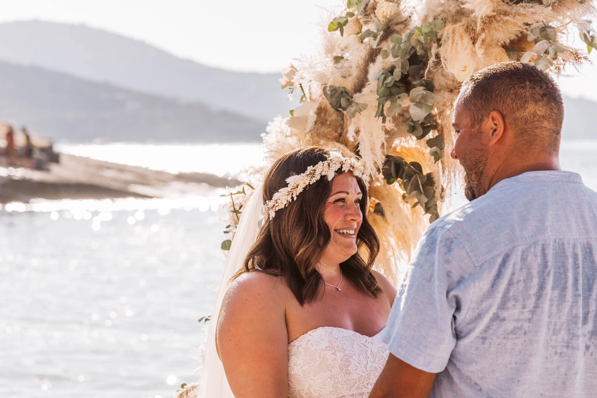 A woman in a white lace wedding dress with a floral crown is smiling at a man in a light blue shirt during an outdoor wedding ceremony by the water, with mountains in the background.
