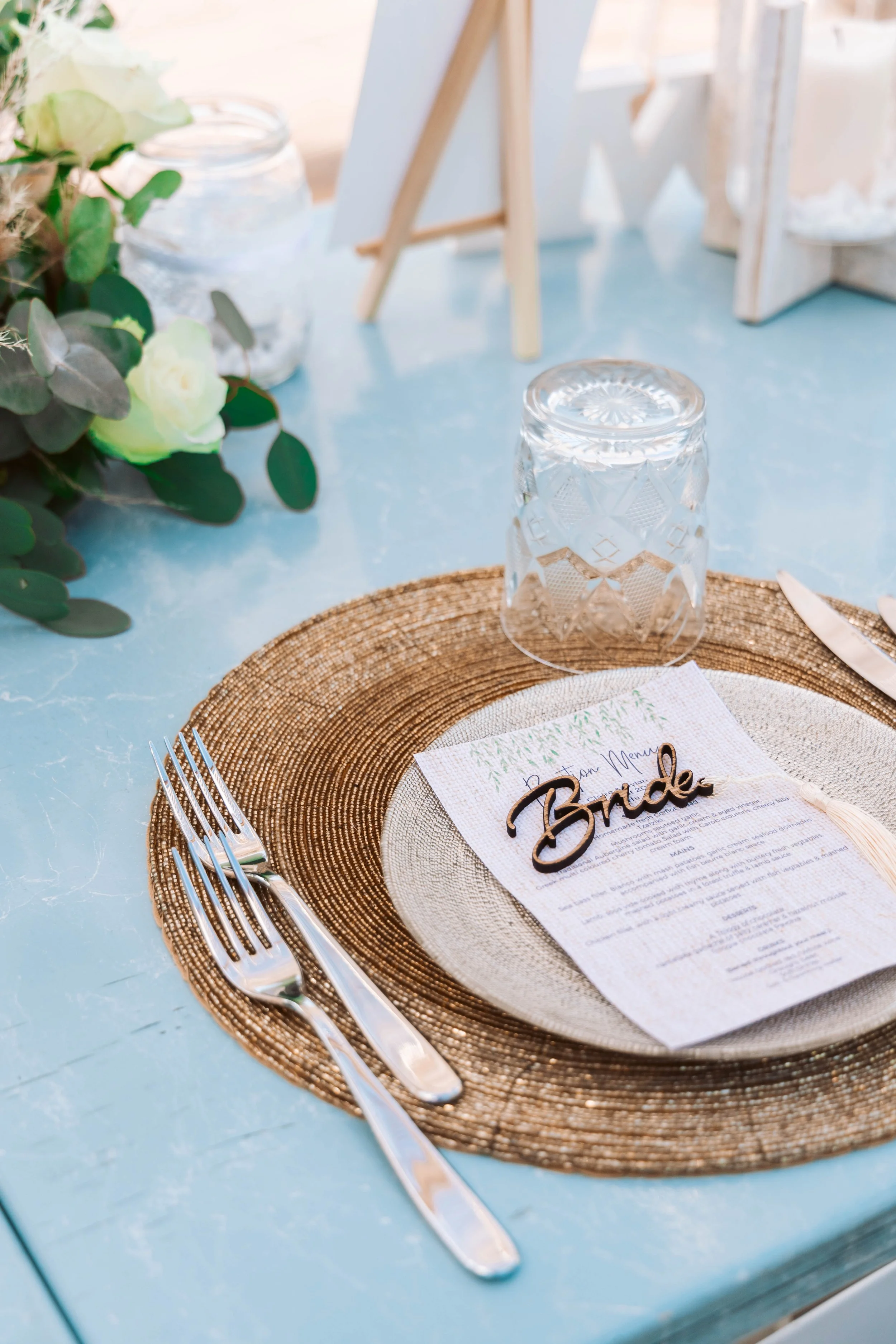 Wedding table setting with a place card labeled 'Bride,' a menu, a gold placemat, water glass, and silverware on a light blue table, decorated with greenery.