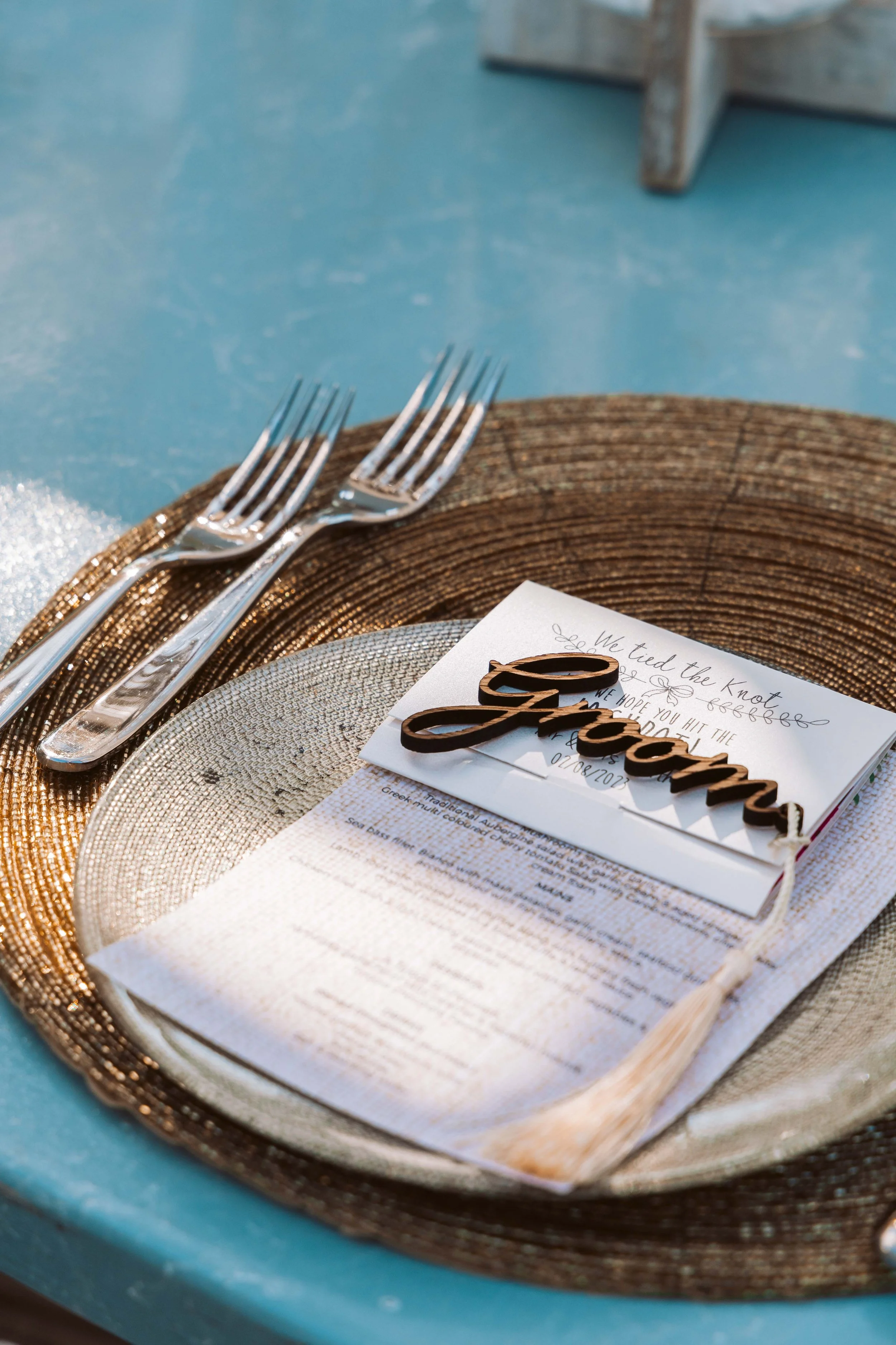 A place setting with a metallic plate, silver utensils, a textured napkin, and a menu card with a decorative 'Groom' sign on top. The table has a turquoise surface, and there is a woven placemat underneath.