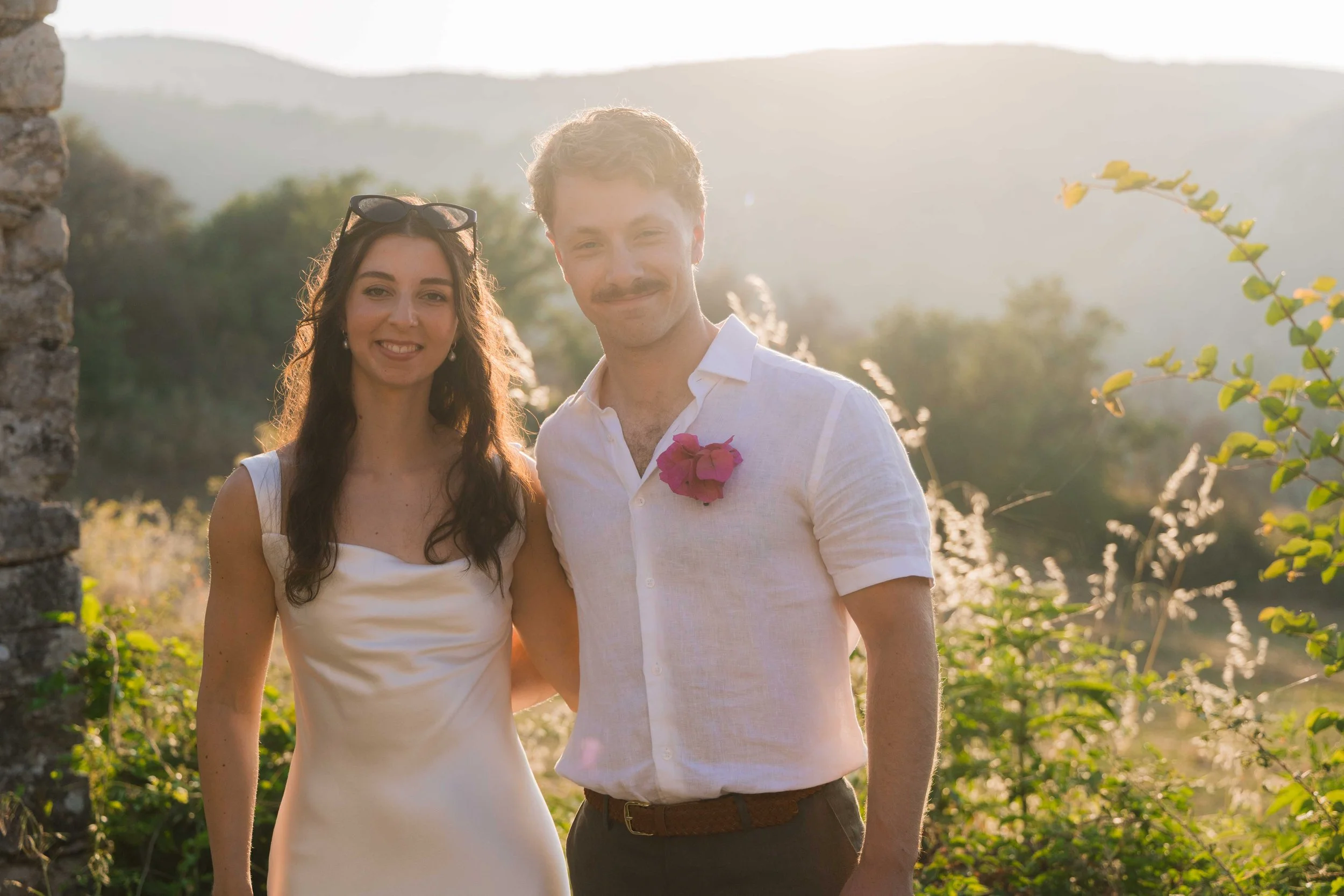 A young couple standing outdoors during sunset, with the woman in a white dress and sunglasses on her head, and the man in a white shirt with a pink flower pinned to it, smiling at the camera.