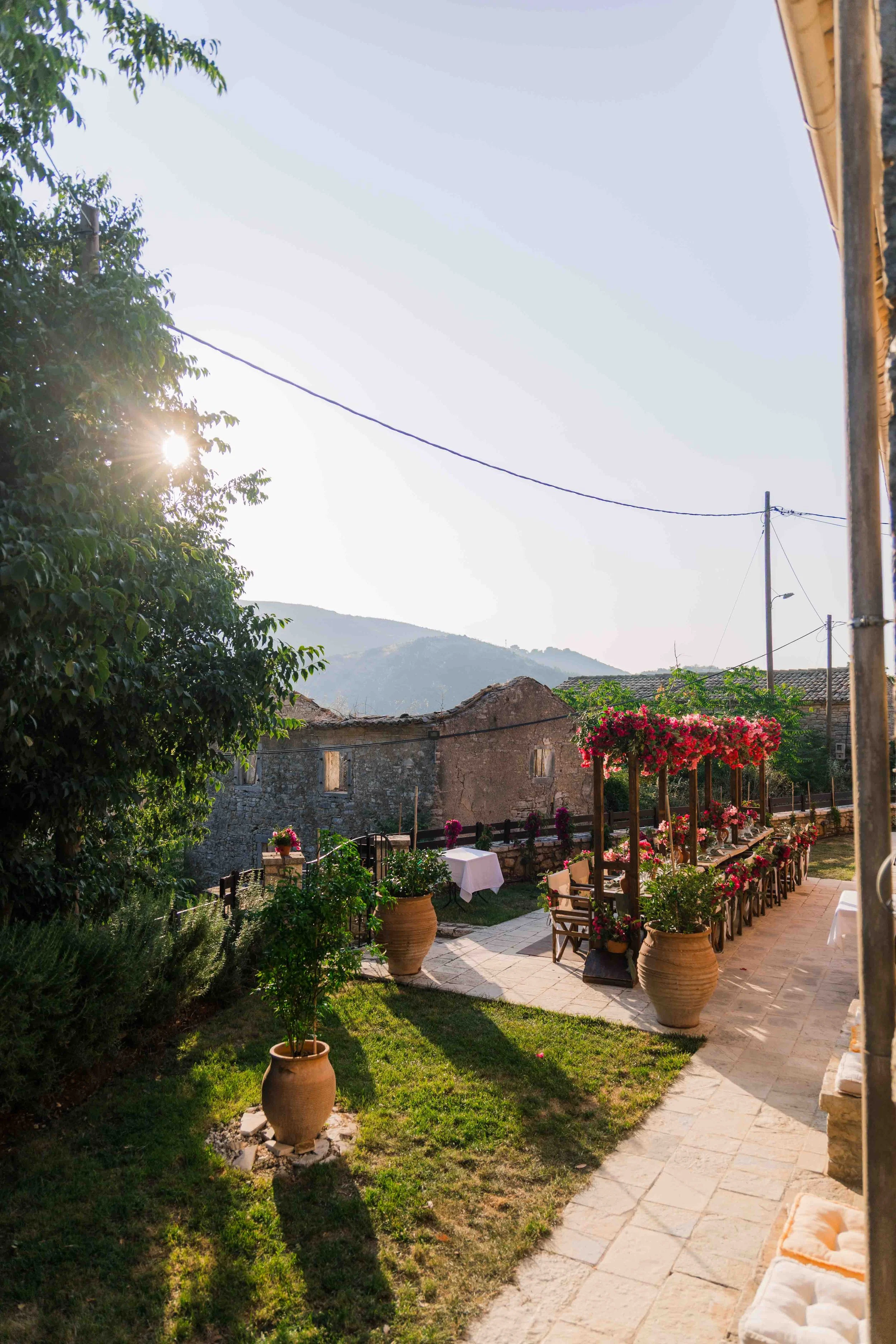 An outdoor patio with picnic tables decorated with pink and red flowers in large pots, surrounded by greenery, under a clear sky with the sun setting behind trees and mountains in the background.